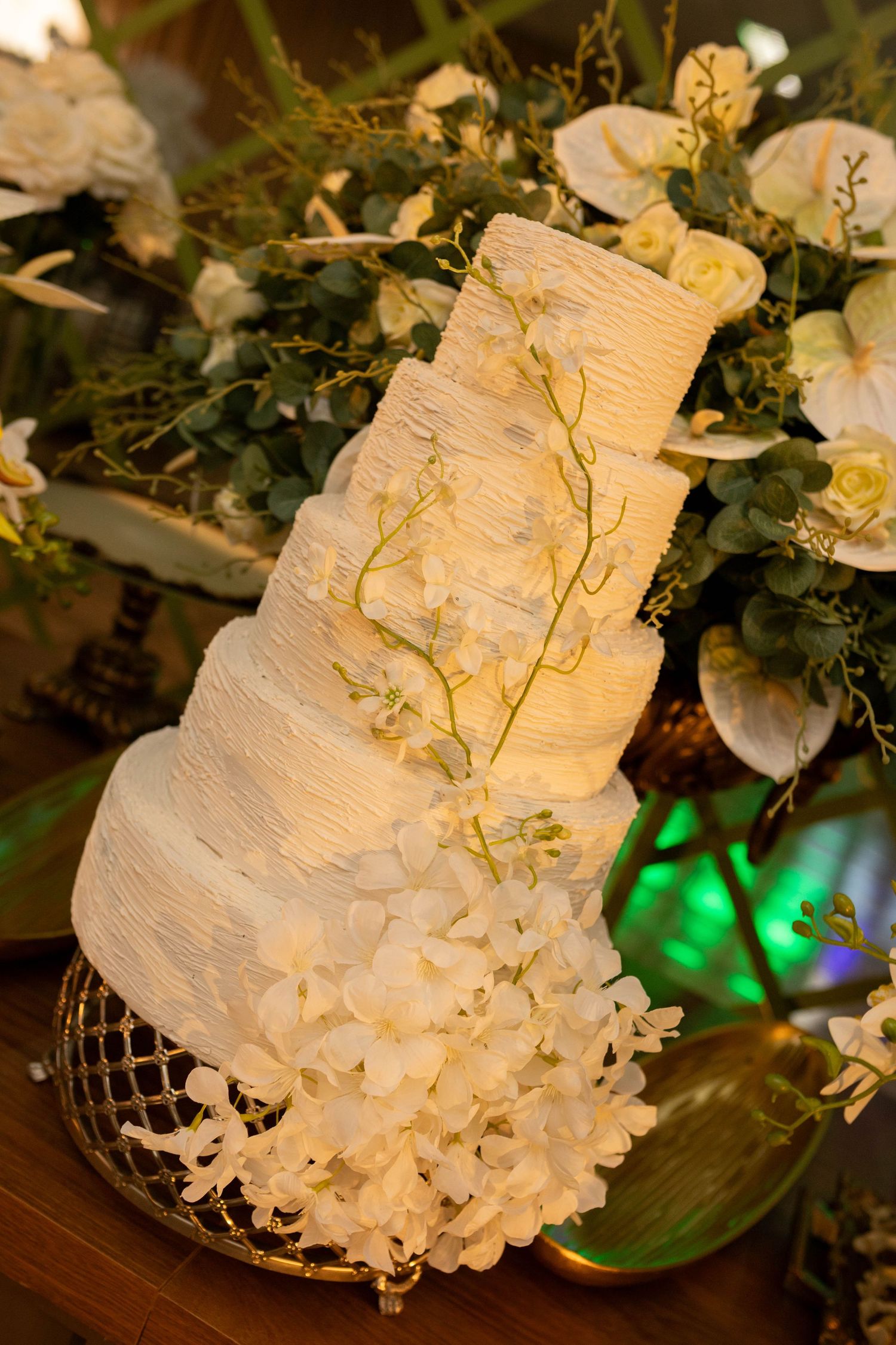 Elegant four-tier white wedding cake adorned with orchids and gold branches on a decorative stand surrounded by lush floral arrangements.