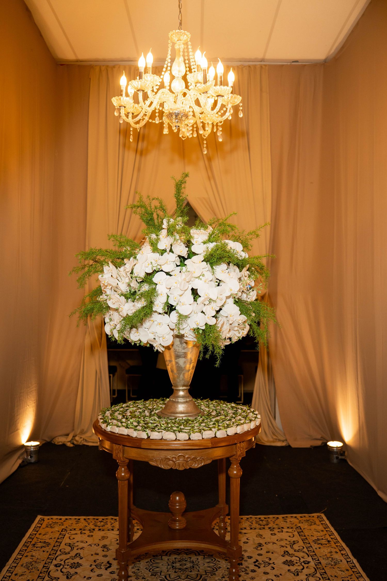 Elegant floral arrangement of white flowers in gold vase on ornate table under crystal chandelier with draped curtains.