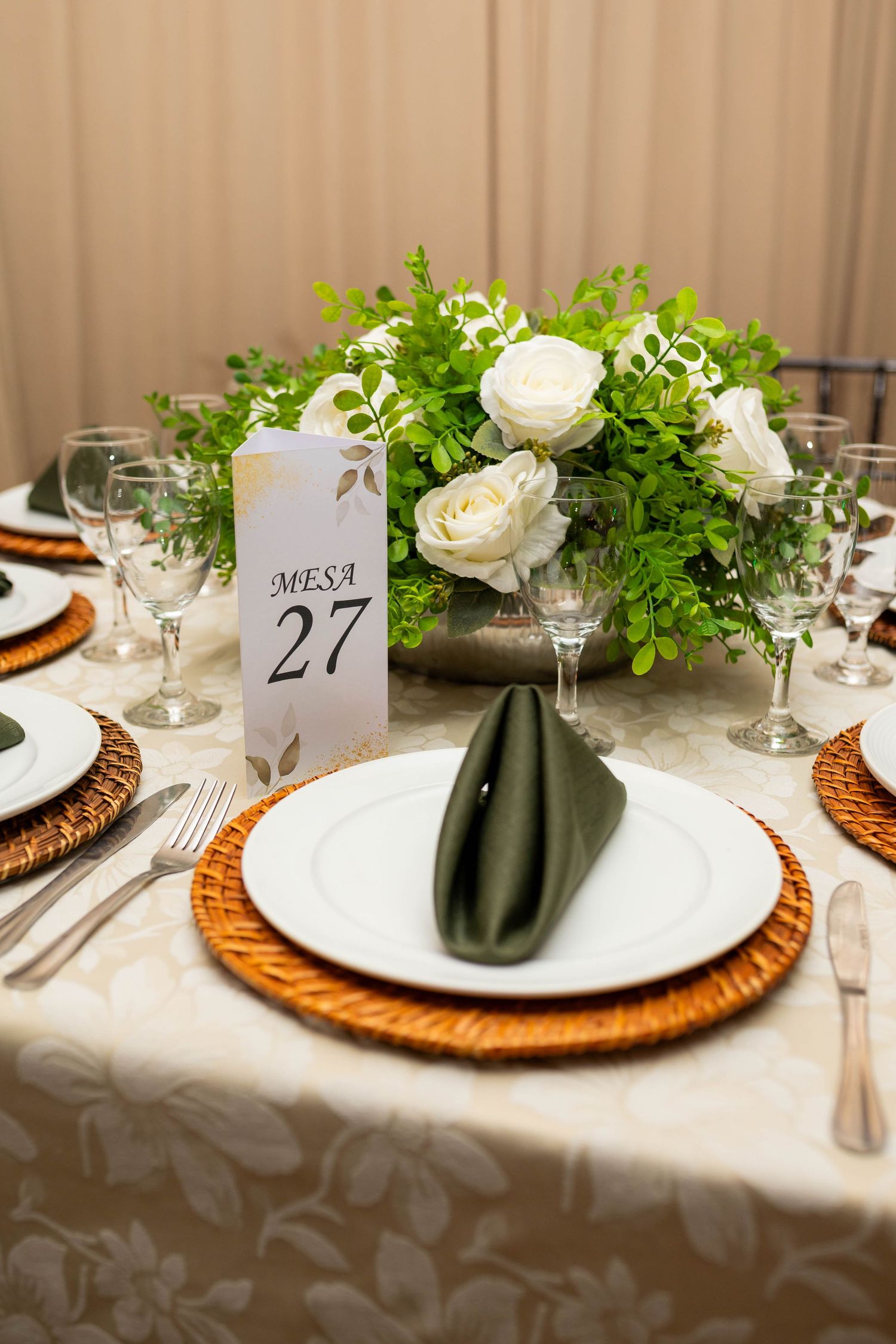 Elegant wedding table setting for Mesa 27 with white roses, greenery centerpiece, wicker chargers, and folded green napkins.