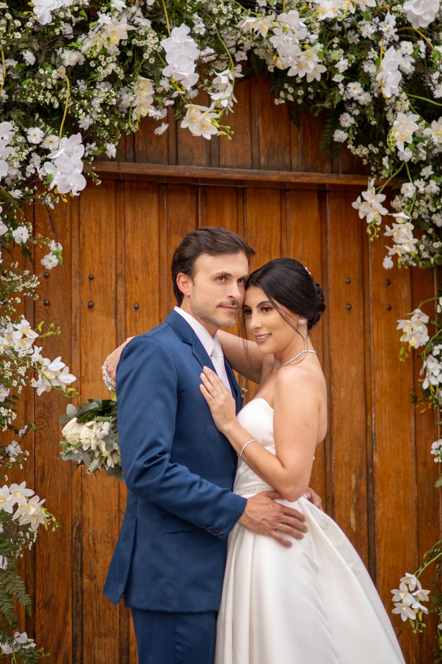 Bride and groom embracing before rustic wooden door adorned with white floral arch on wedding day.