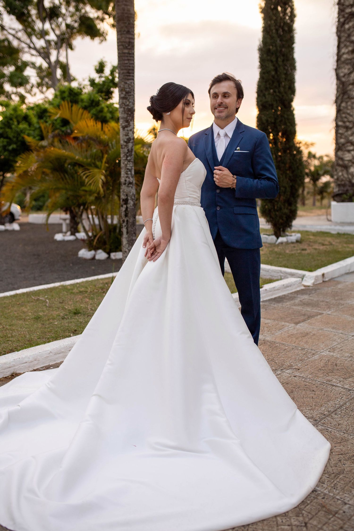 Bride in white gown with long train stands with groom in navy suit outdoors at sunset wedding venue.