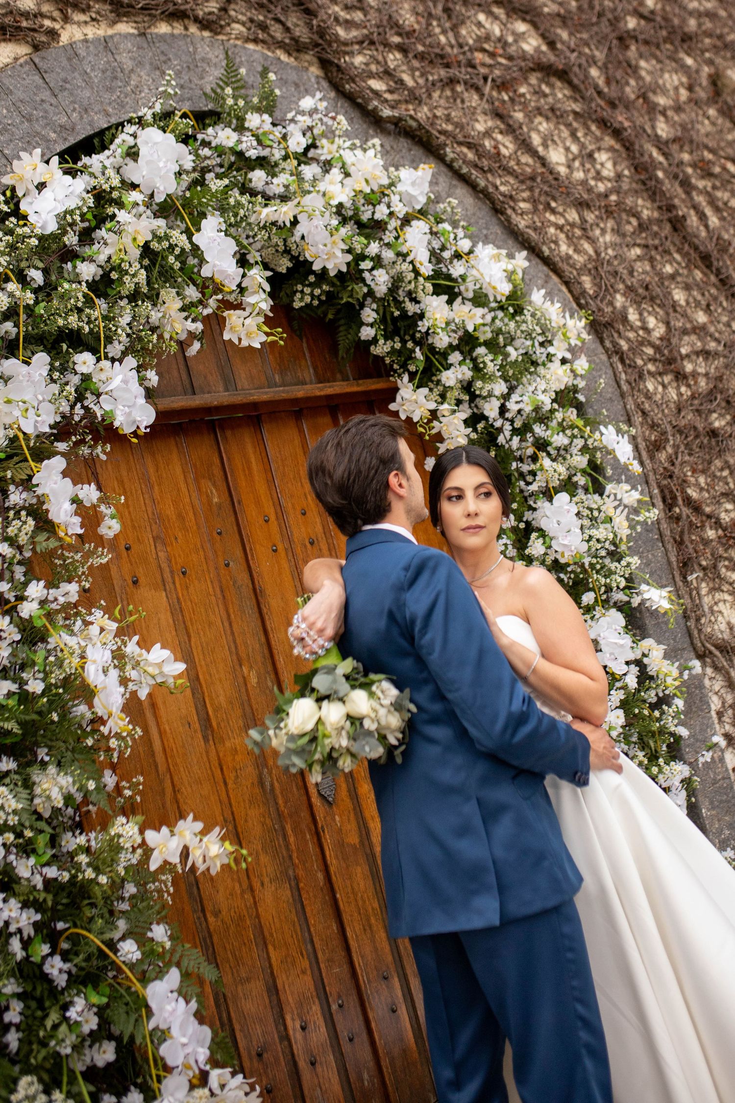 Bride and groom embrace before a wooden door adorned with a white floral arch at their wedding ceremony.