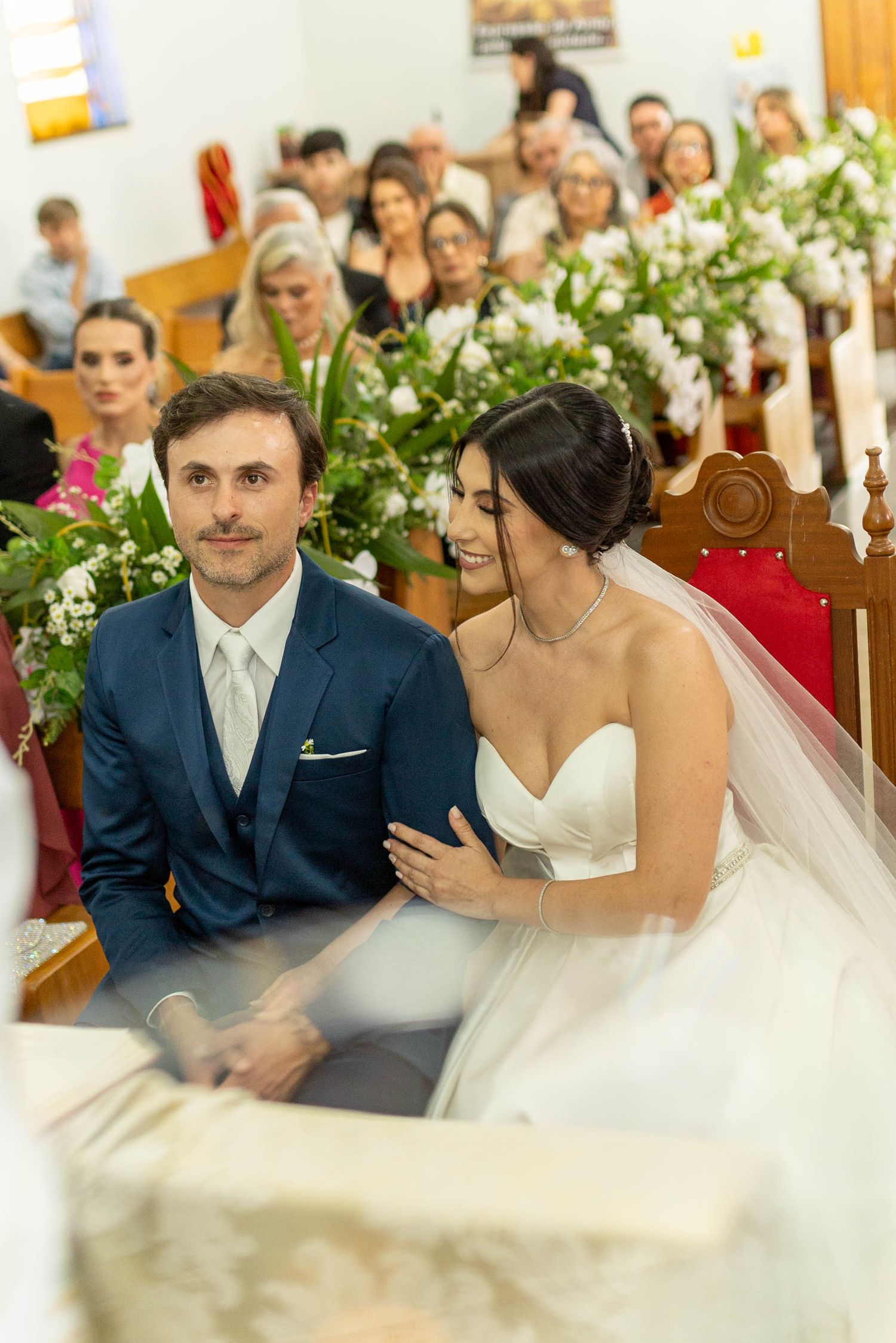 Bride and groom sitting together at church altar during wedding ceremony, guests seated in pews behind them.