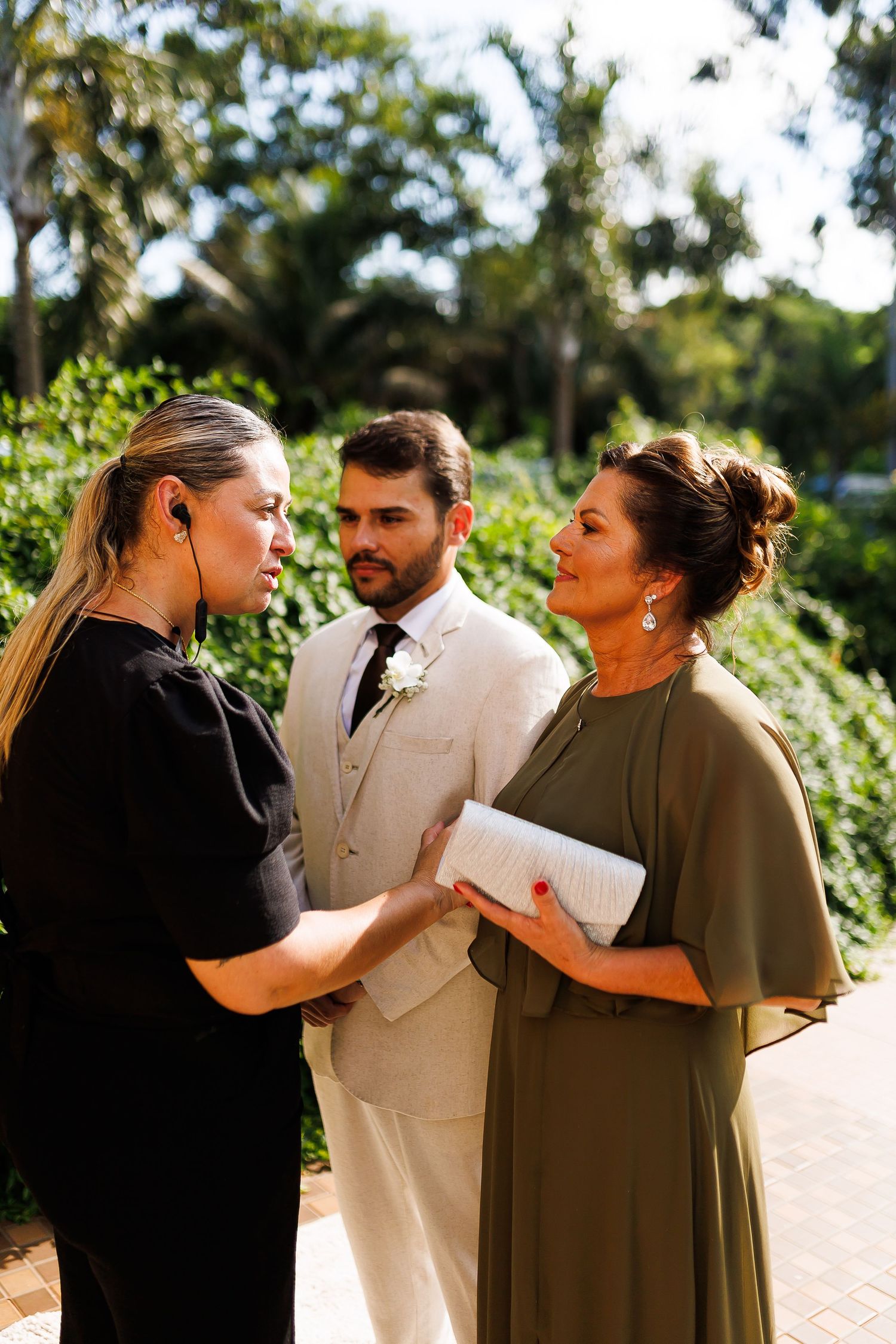 A woman in black dress holds hands with woman in olive gown as a groom in white suit stands behind them outdoors.