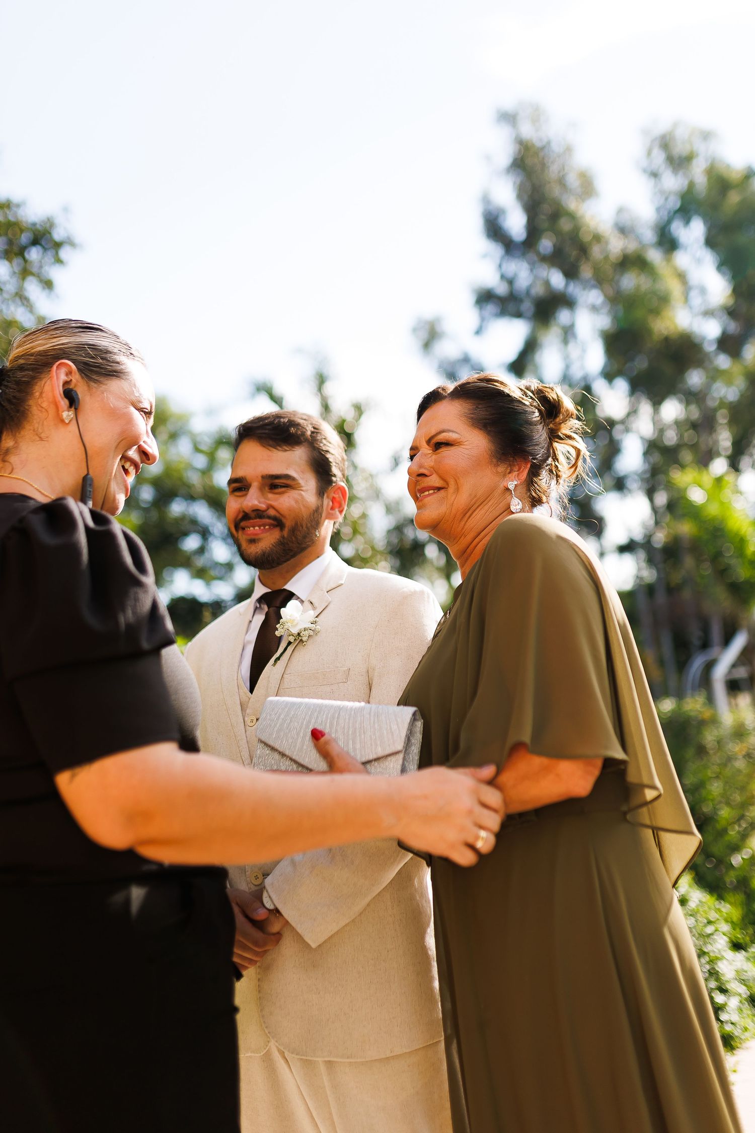 Groom in beige suit with boutonniere smiles during outdoor wedding ceremony with two women in black and olive dresses.
