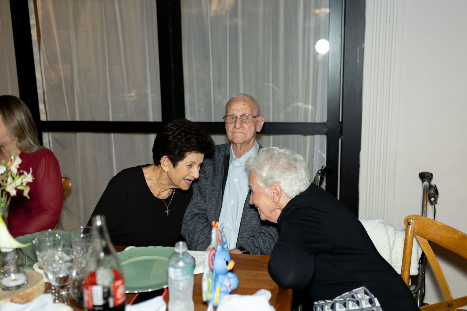 Three elderly people engaged in conversation at a dining table during an indoor gathering or dinner event.