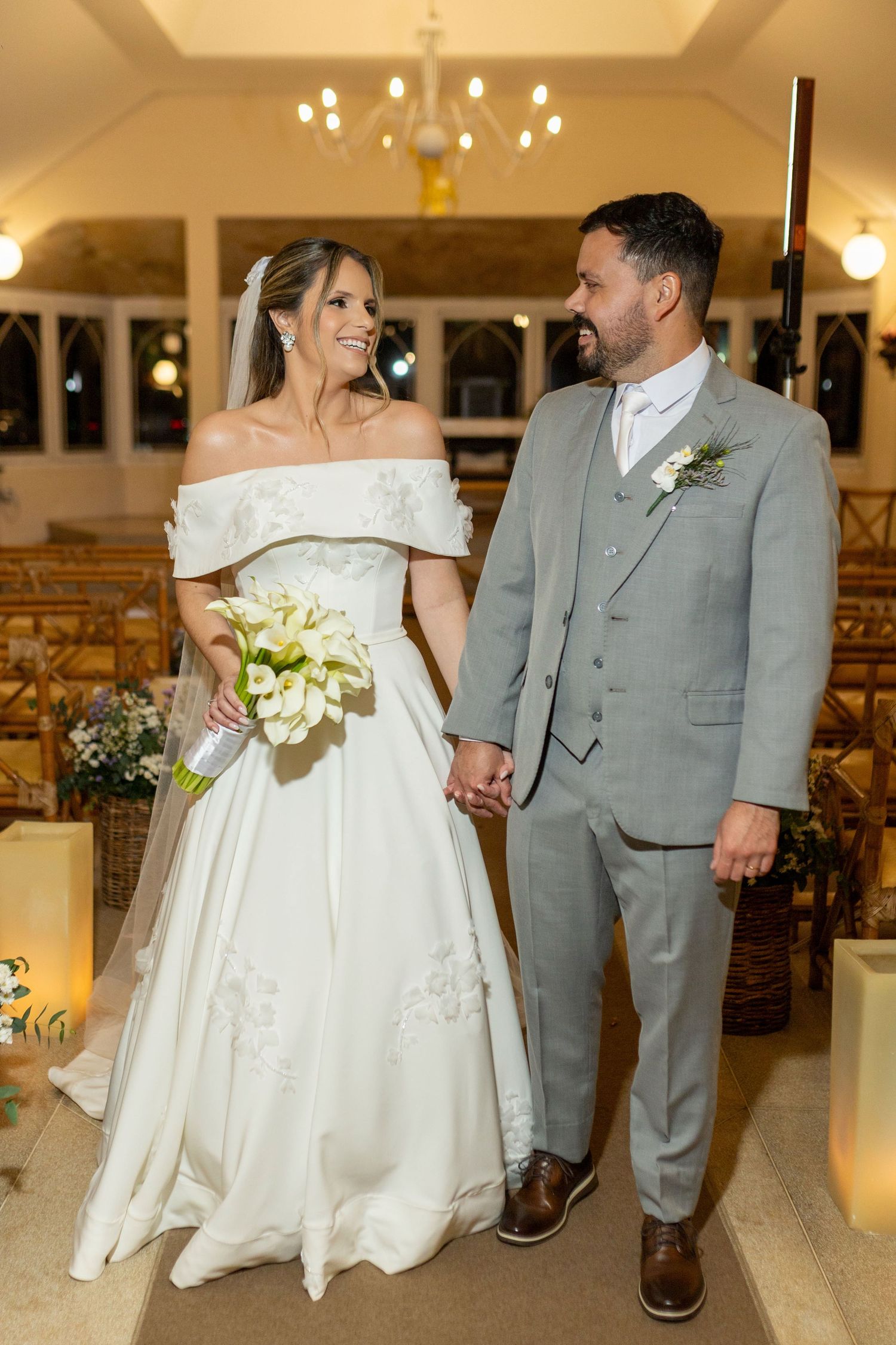 Bride in off-shoulder white gown holding calla lilies and groom in gray suit holding hands at church wedding ceremony.