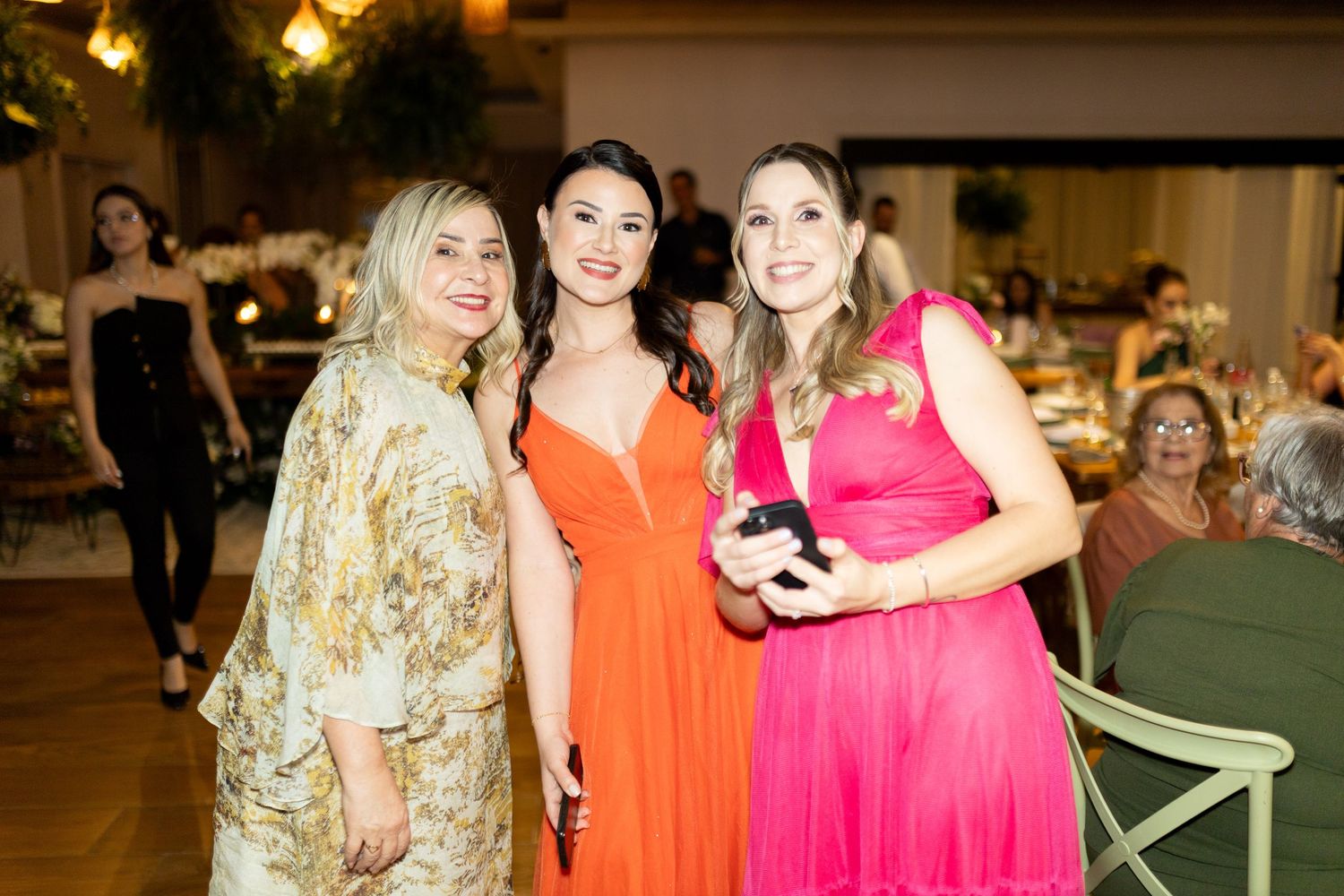 Three elegantly dressed women pose together at a formal event, wearing colorful gowns in floral, orange, and pink.