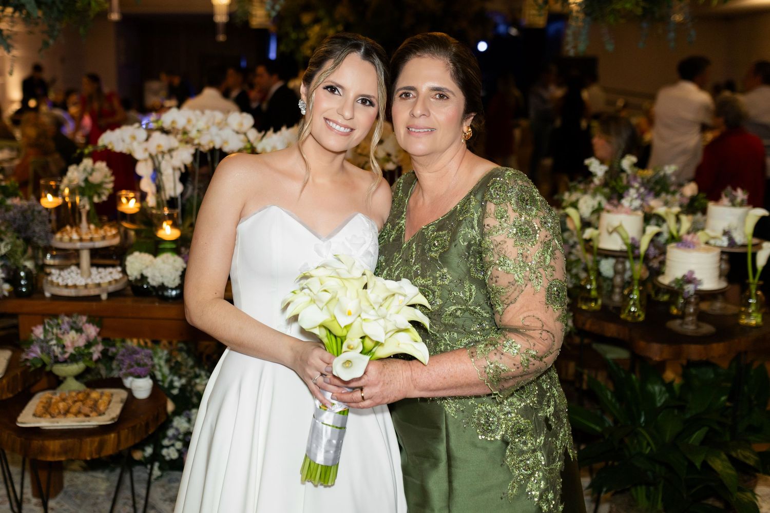 Bride in white strapless gown holding white calla lily bouquet poses with woman in green dress at elegant wedding reception.
