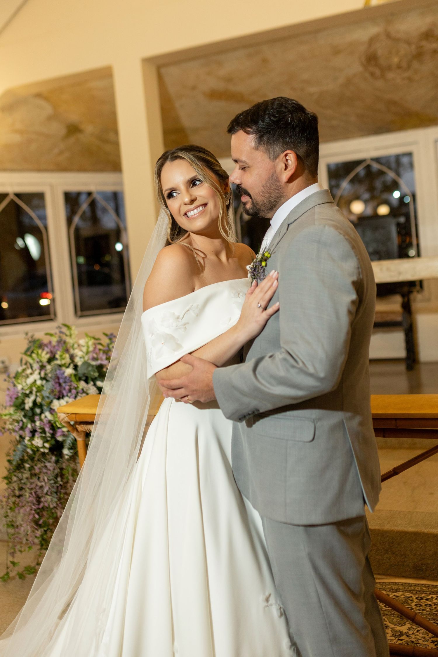 Bride in white off-shoulder gown and groom in gray suit share a romantic embrace in a decorated wedding venue.