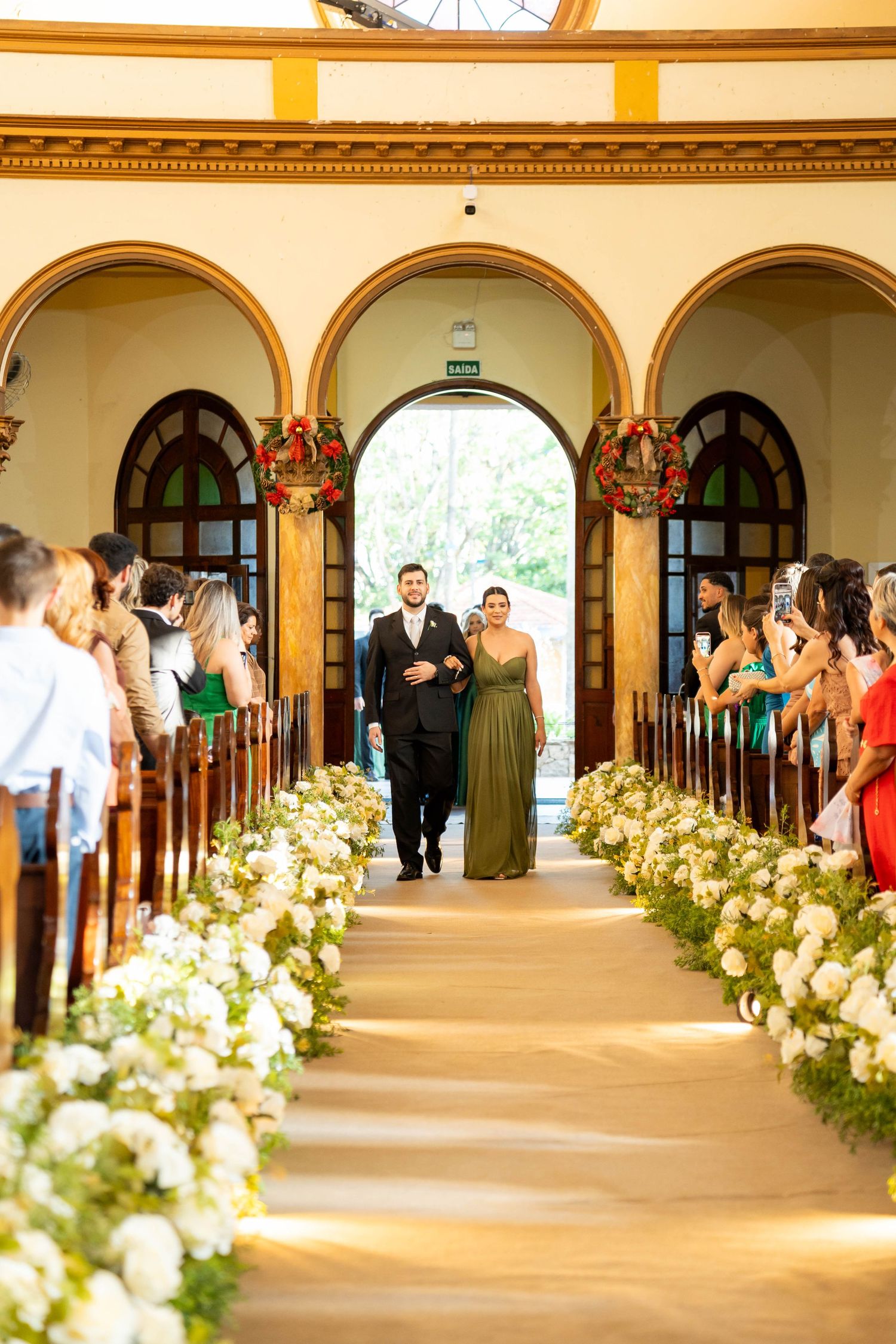 Elegant wedding ceremony aisle lined with white flowers, couple walking toward altar in a beautiful arched chapel.