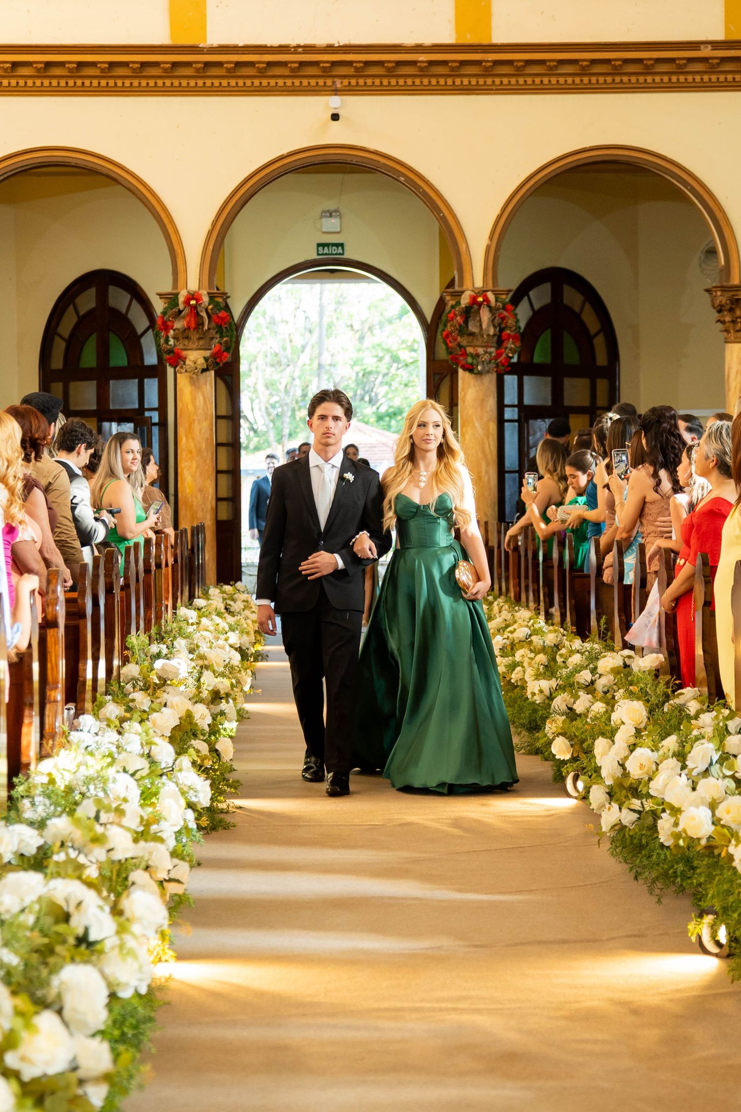 A couple walks down a flower-lined church aisle, the woman in an emerald green gown and the man in a black tuxedo.
