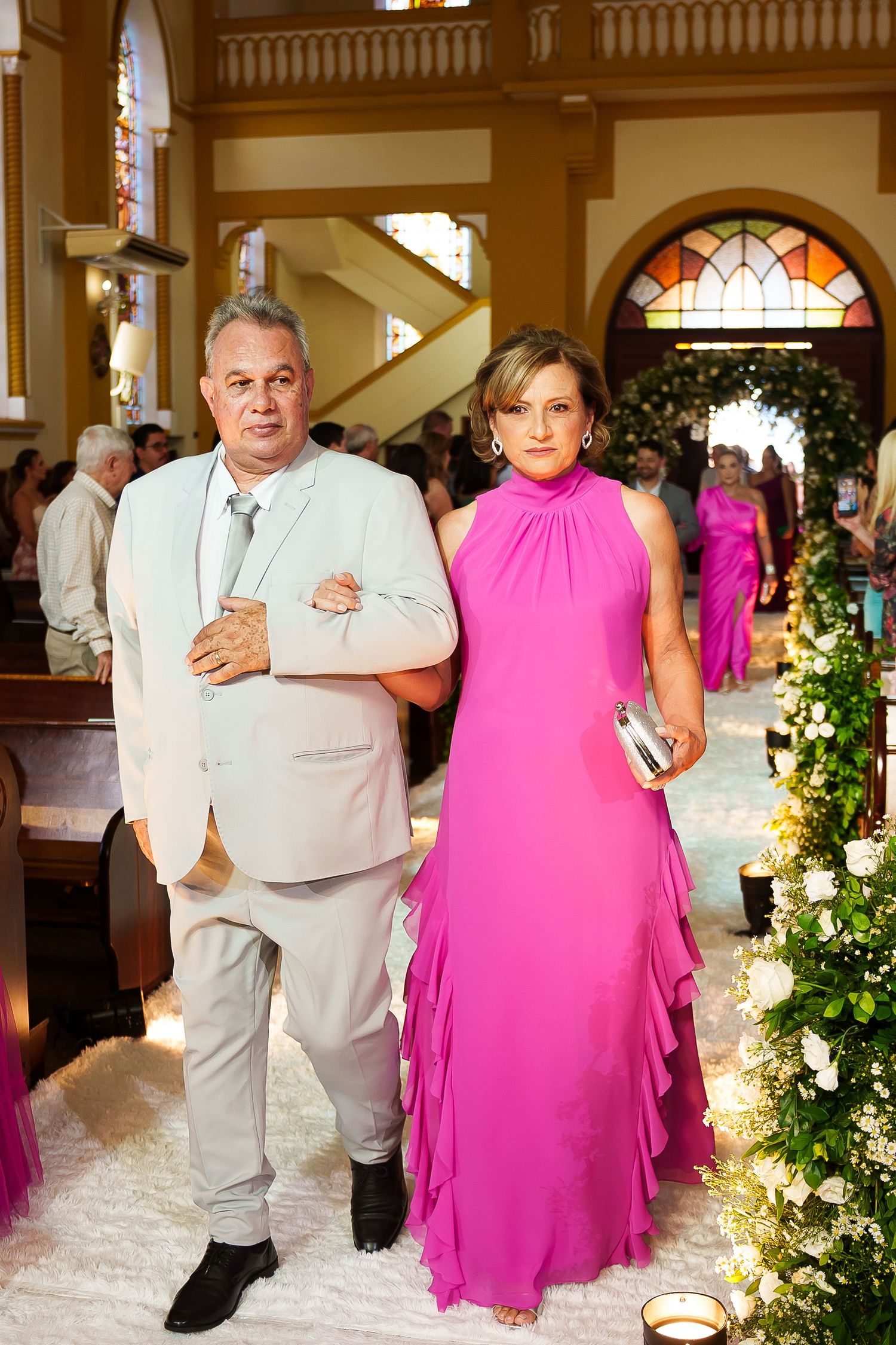 Elegant couple posing in a church, man in light suit, woman in vibrant pink halter gown, floral aisle decorations.