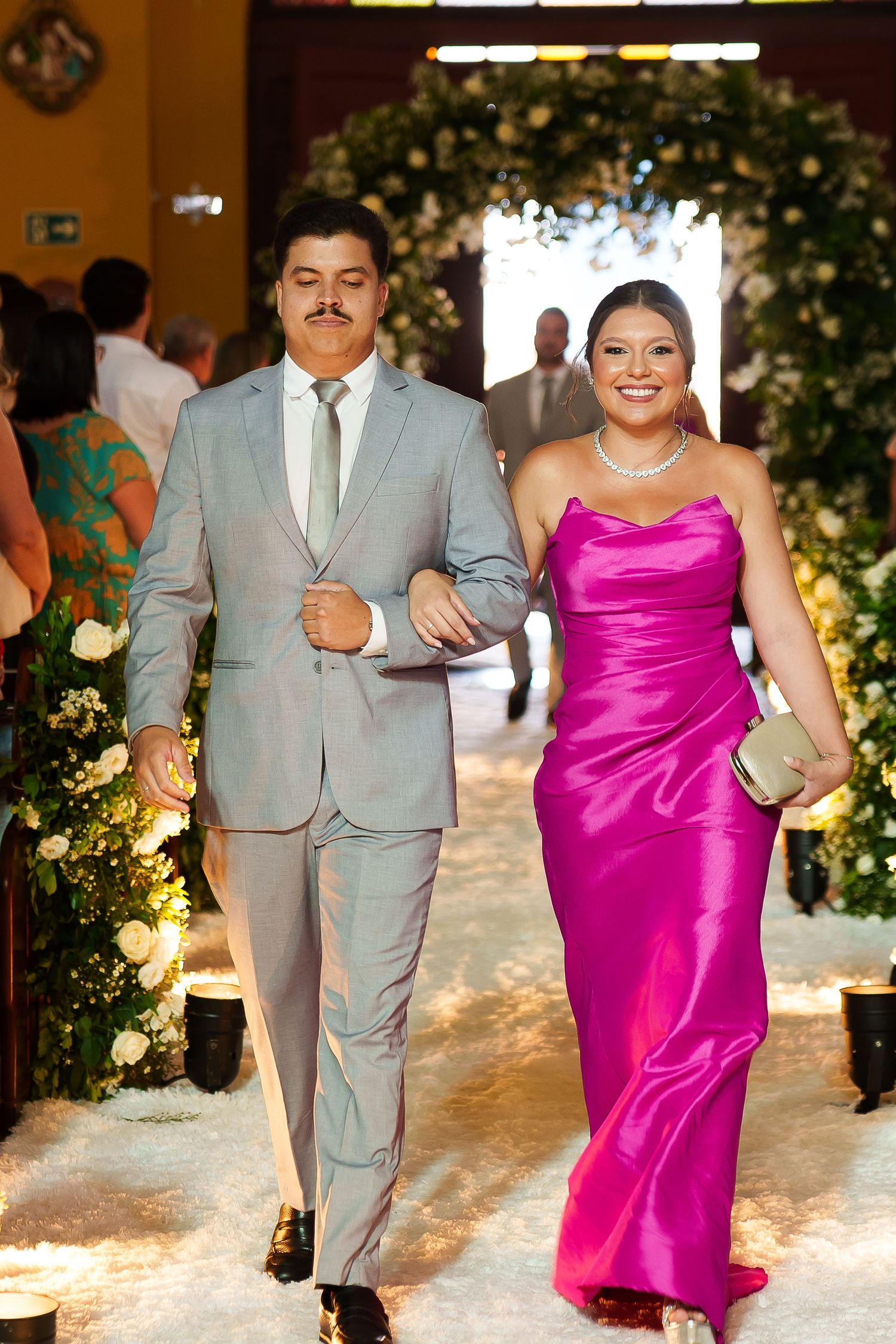 Couple walking down wedding aisle, man in gray suit, woman in magenta satin gown, floral decorations.