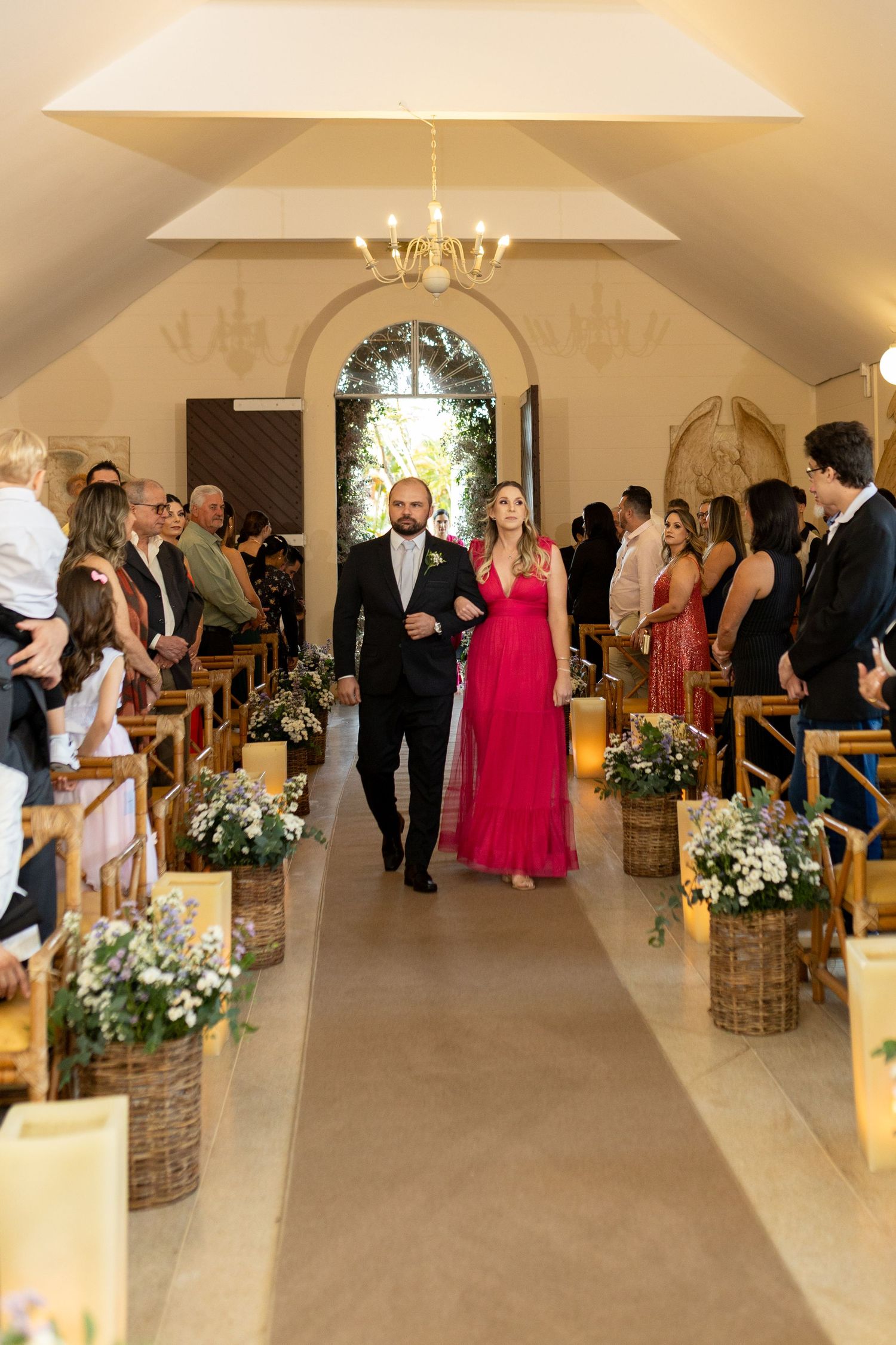 Couple walks down chapel aisle during wedding ceremony with floral decorations and guests seated.