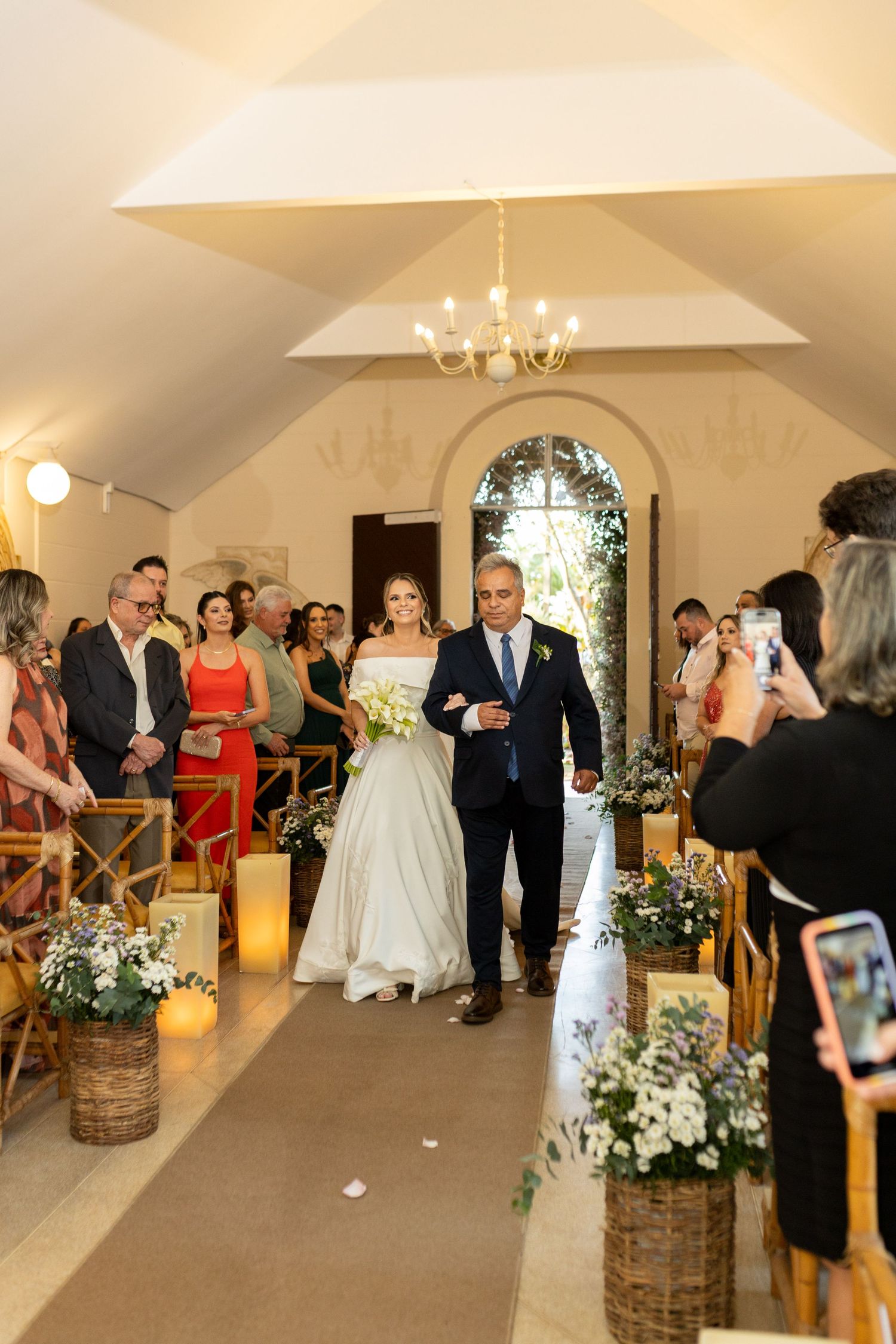 Bride walks down the aisle with escort in elegant chapel wedding ceremony, guests standing, floral decorations lining the path.