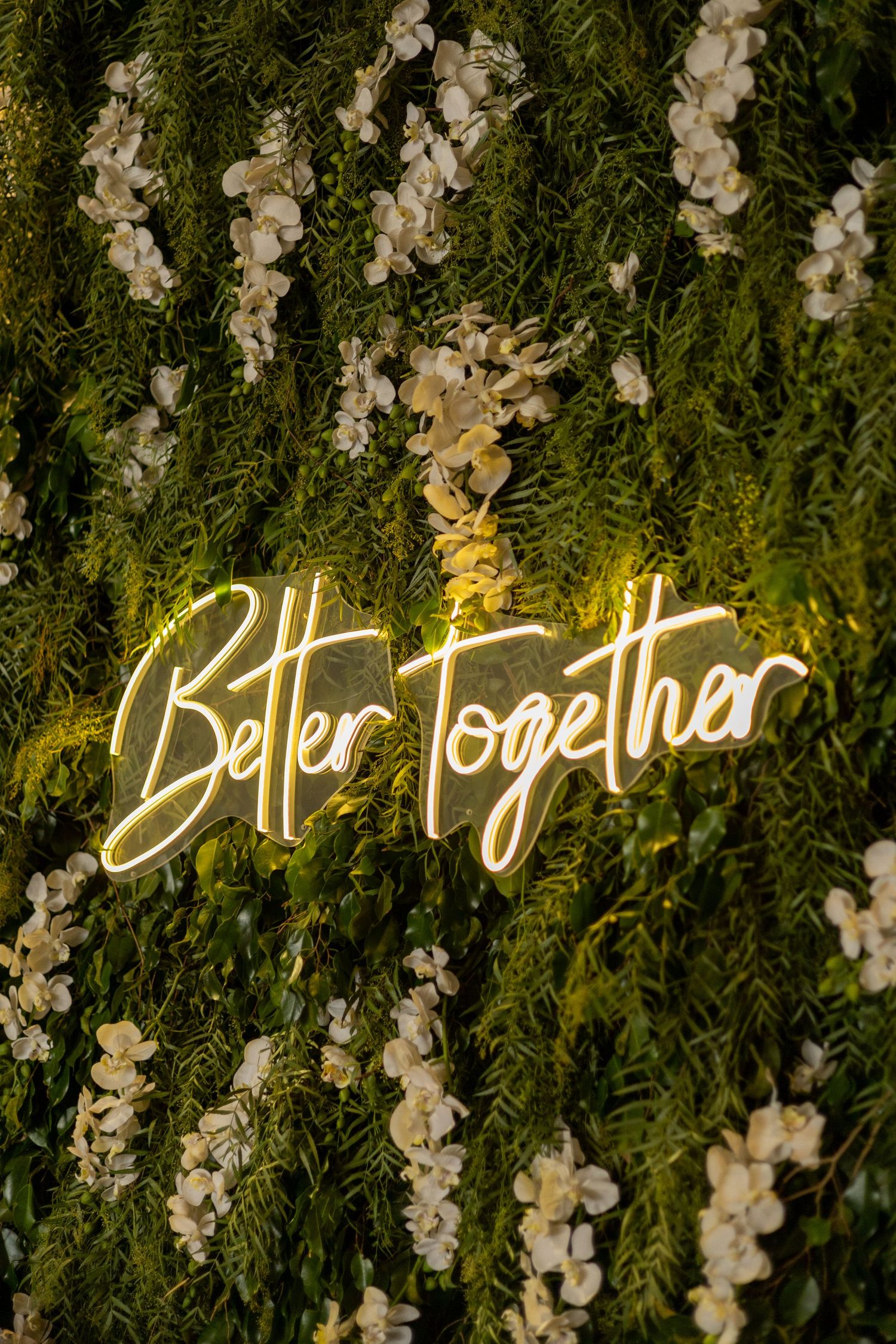 Glowing neon sign reading Better Together on a lush green floral wall with white orchids at a wedding.