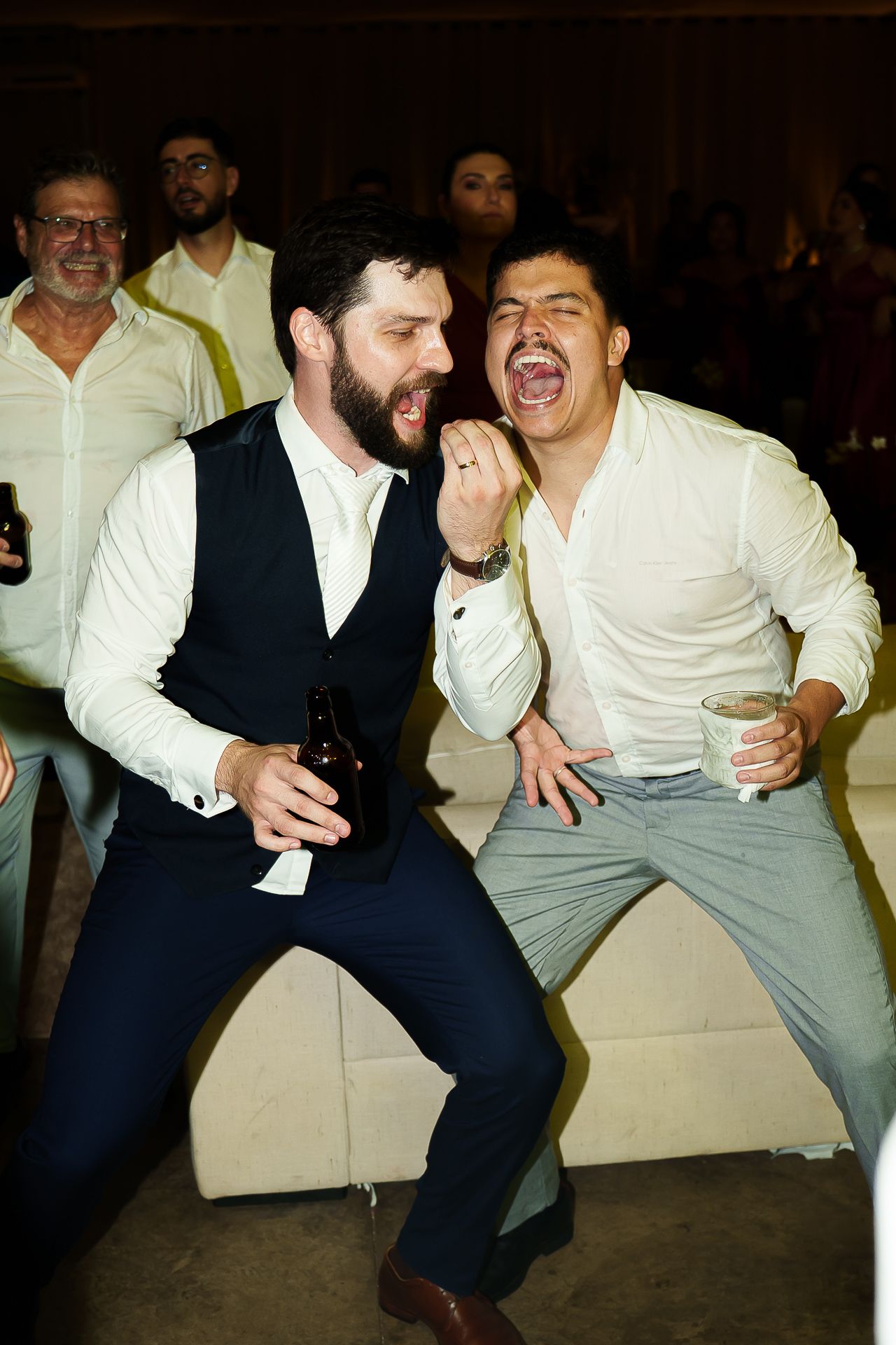 Two men in white shirts laugh and dance together at a party, one holding a beer bottle and the other a cup.