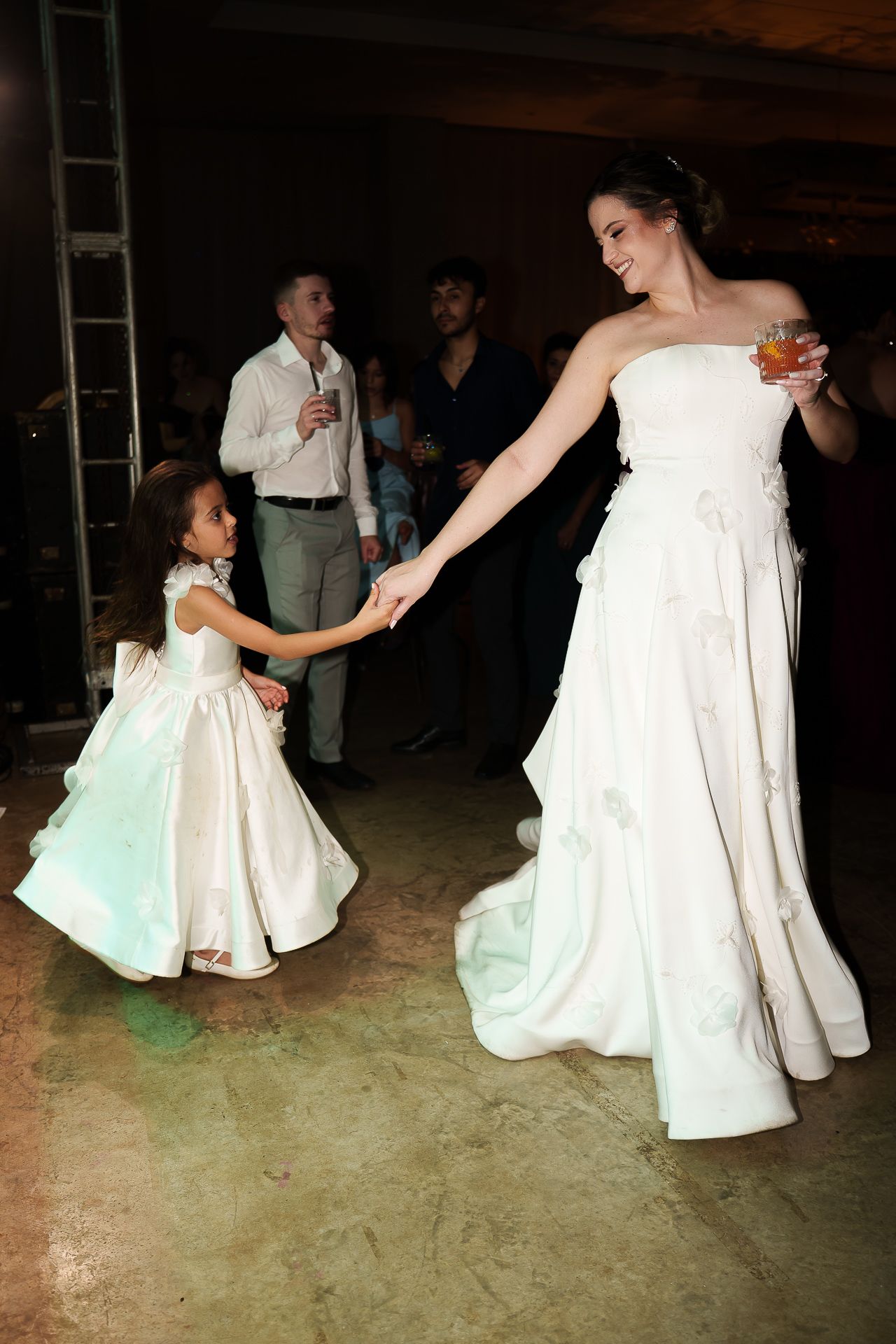 Bride in white gown dancing with flower girl at wedding reception while guests watch in background.