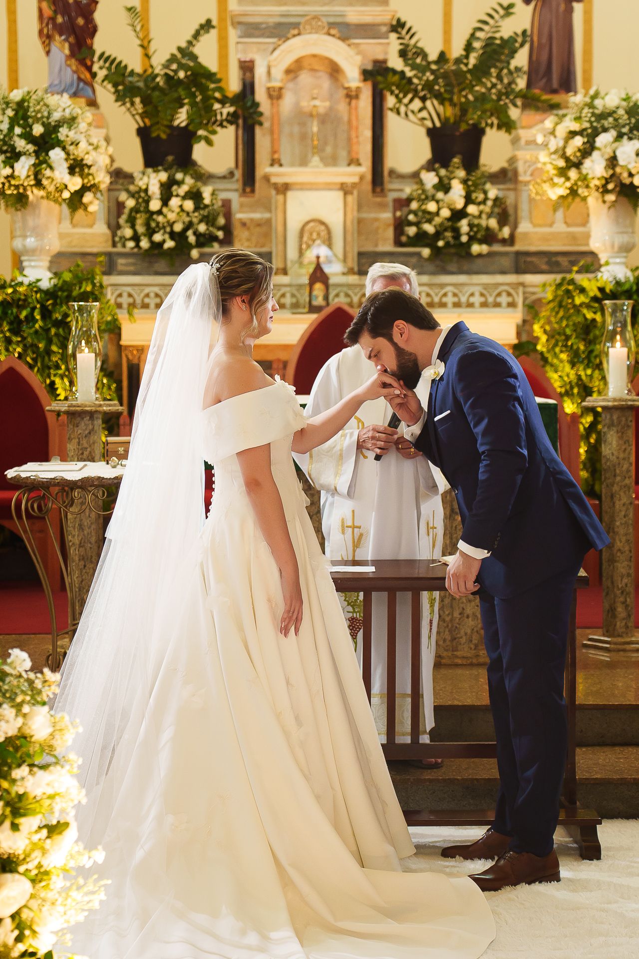 Groom in blue suit kissing bride's hand at church altar during wedding ceremony with priest and floral decor.