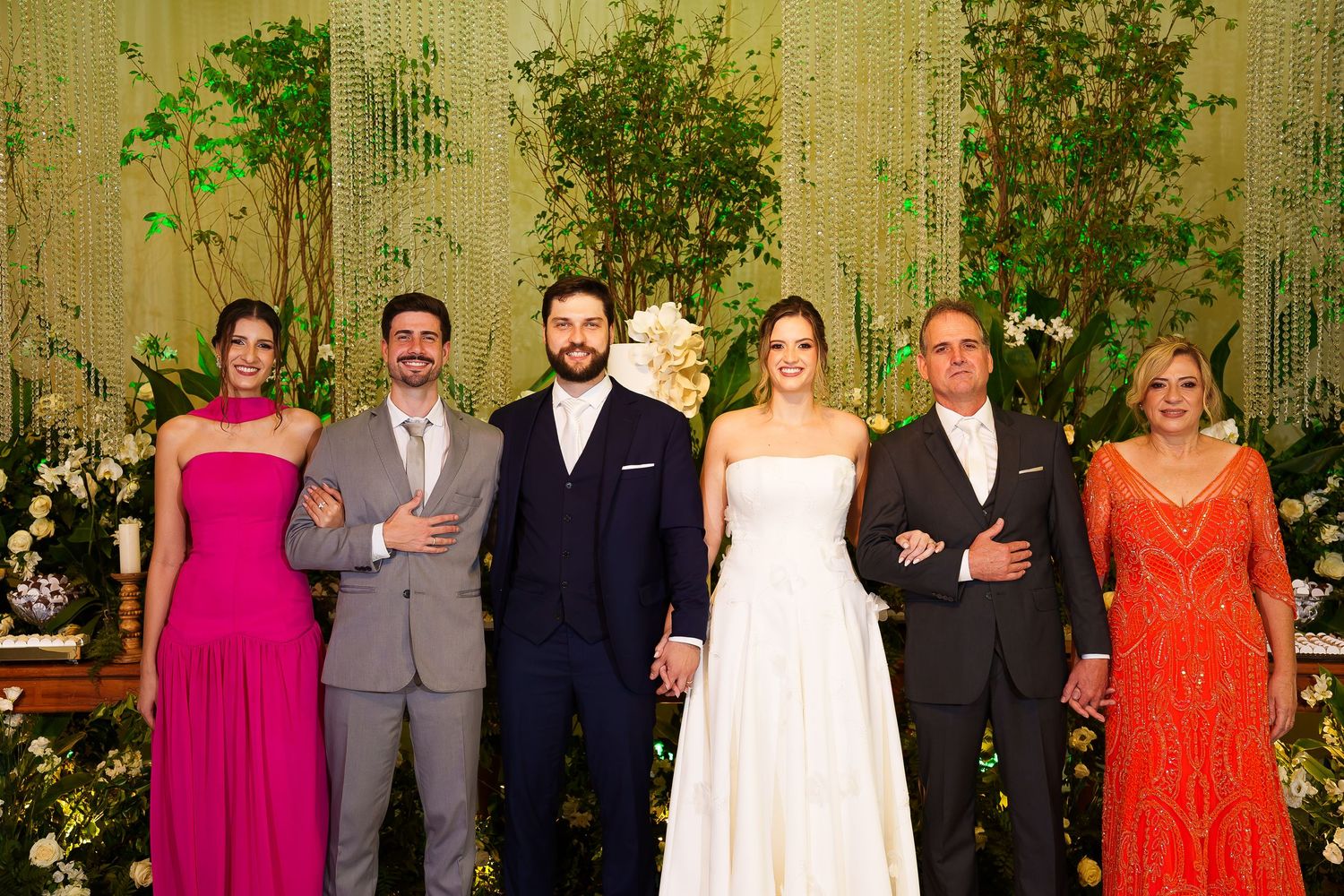 Wedding party of six posing together, bride in white gown, guests in formal attire, floral backdrop.