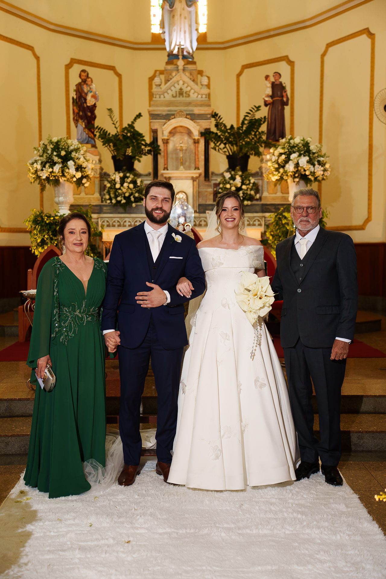 Bride and groom pose with family members at church altar, bride in white gown holding bouquet, guests in formal attire.