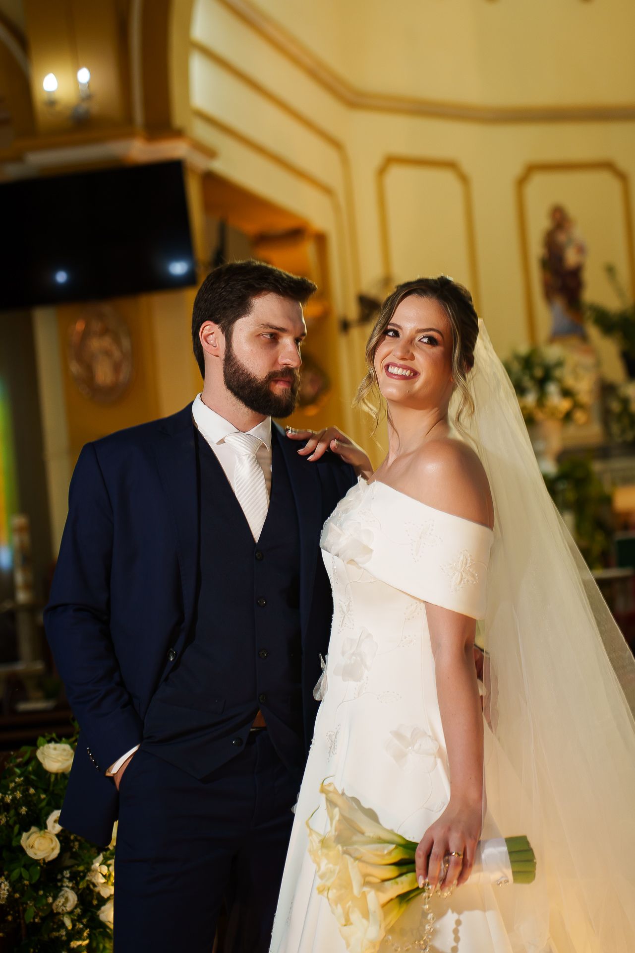 Bride and groom pose elegantly in a church, she in an off-shoulder white gown holding yellow roses, he in a navy suit.