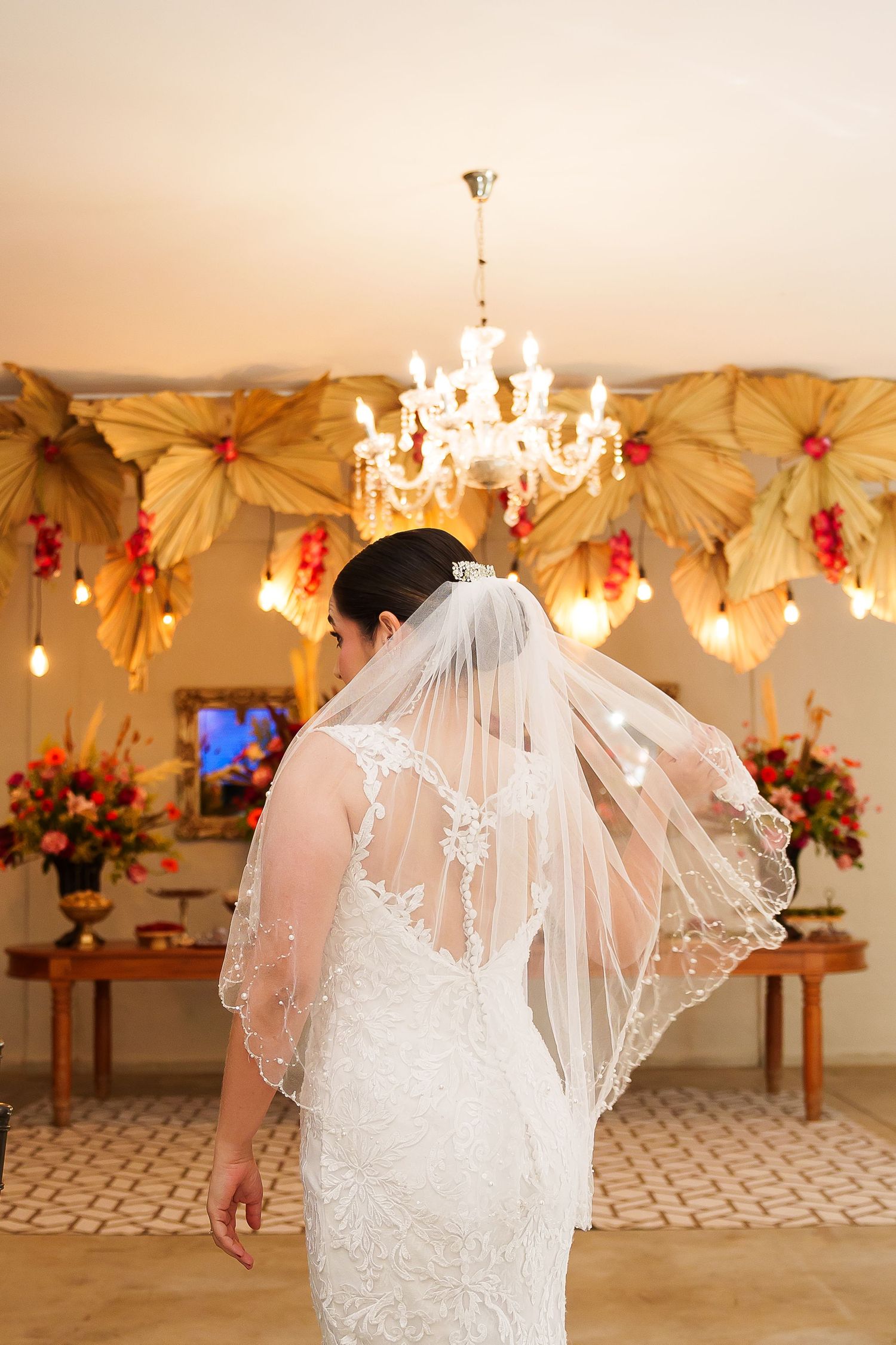 Bride in lace wedding dress and veil stands before chandelier and floral decor at elegant wedding venue.