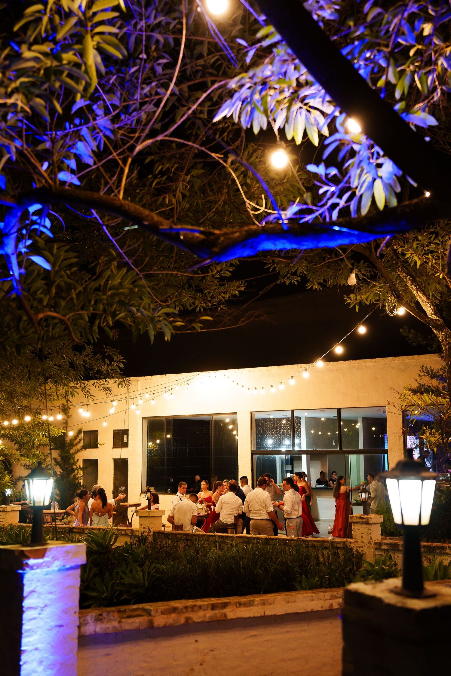 Outdoor evening wedding reception under string lights and trees with guests mingling near a warmly lit venue.