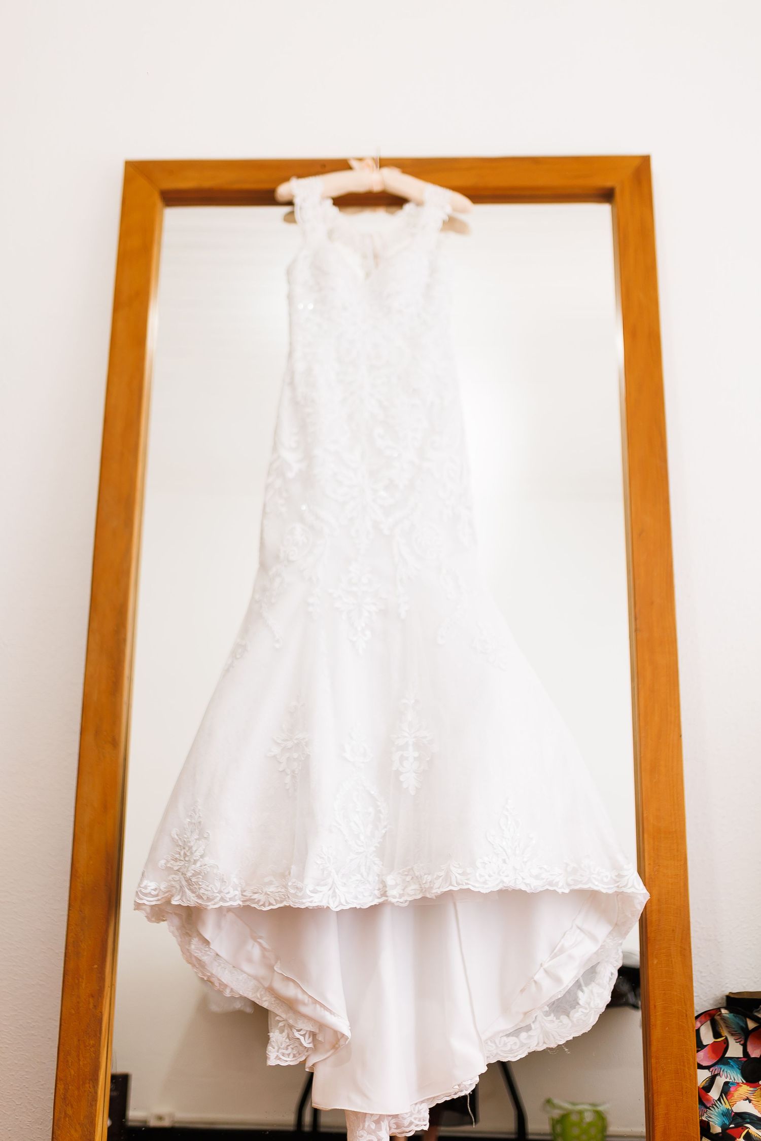 Elegant white lace mermaid wedding dress hanging on a wooden mirror frame against a white wall.
