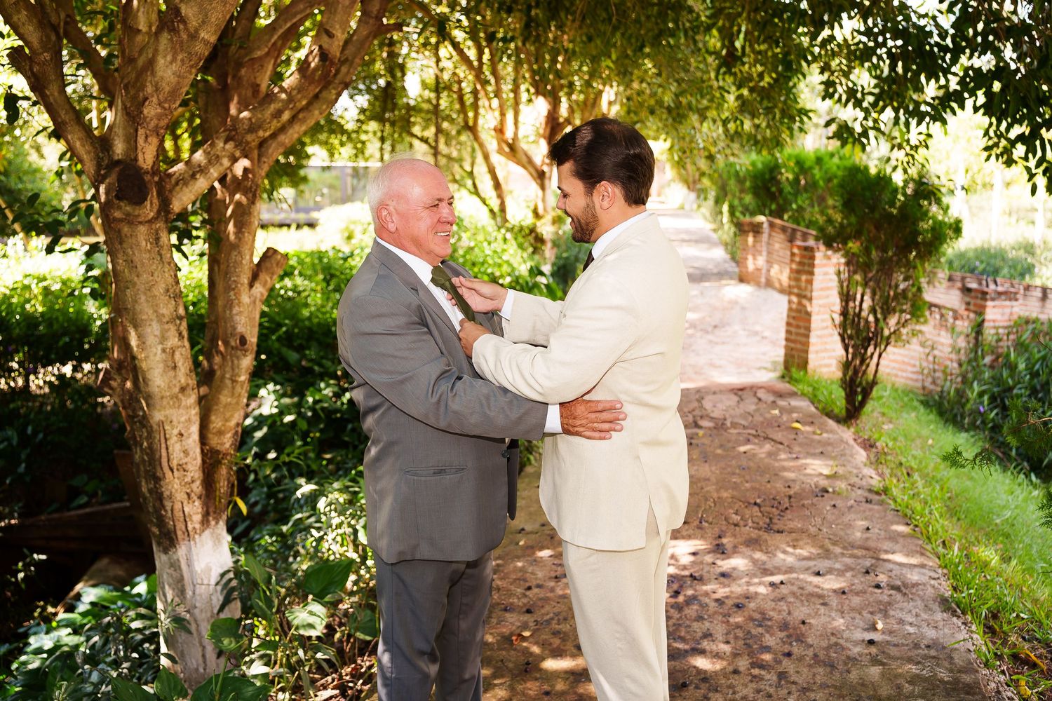 Two men in suits share a tender moment outdoors, adjusting each other's attire near trees at a wedding.