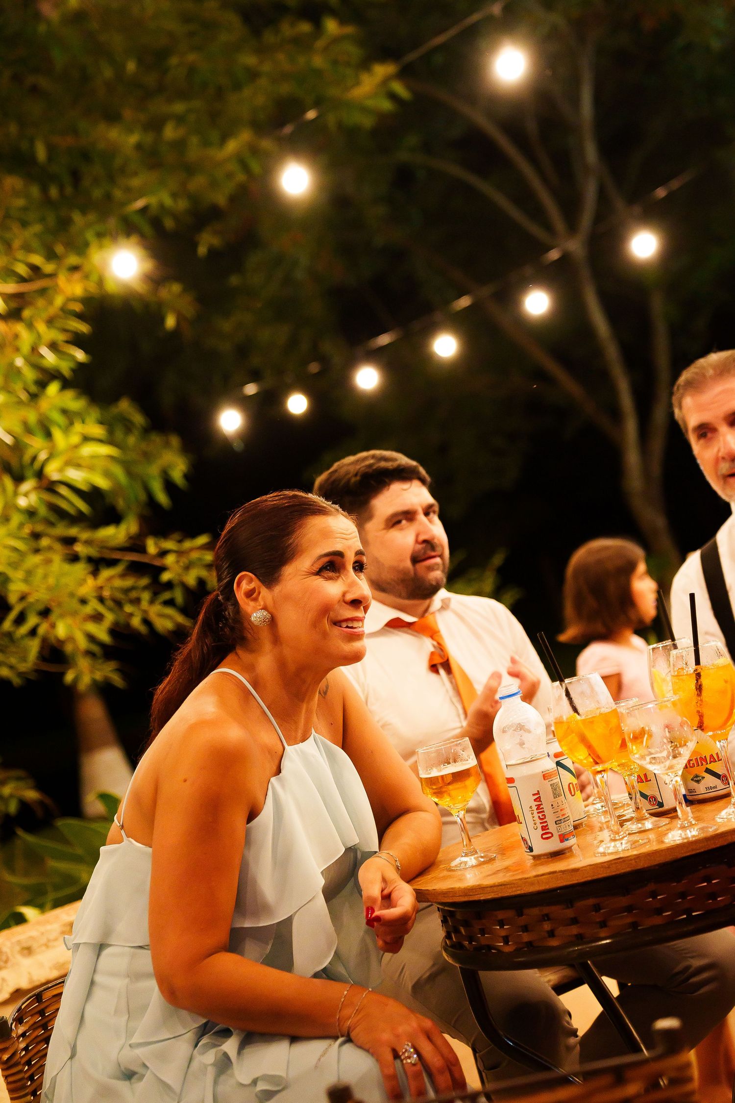 Guests seated at outdoor evening wedding reception table with string lights glowing among trees in background.