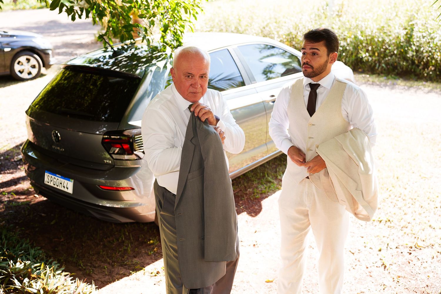 Two men in formal attire stand outdoors near a car, one helping the other put on a suit jacket.