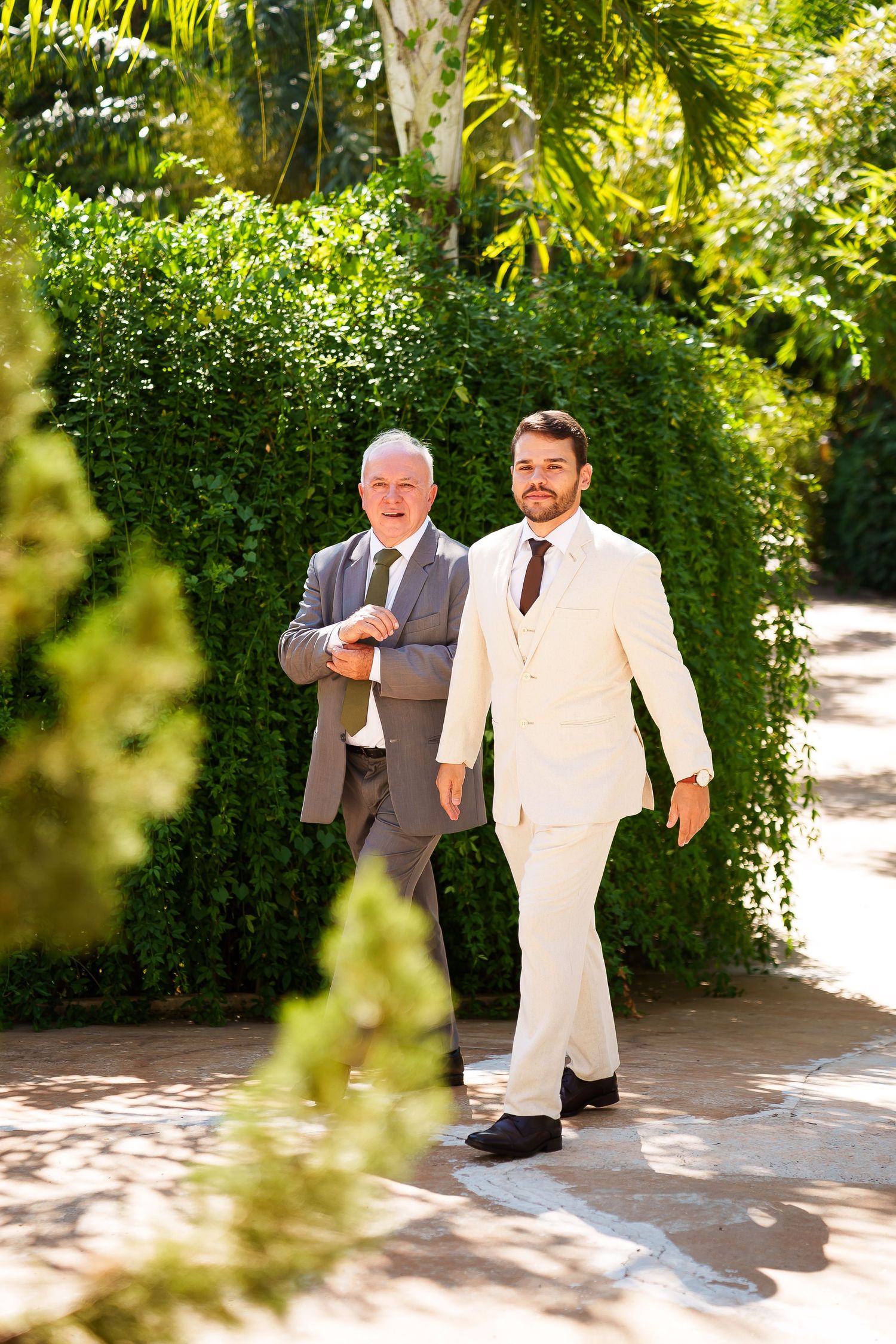 Two men in suits walk through a lush garden, one in cream and one in gray, likely at a wedding ceremony.