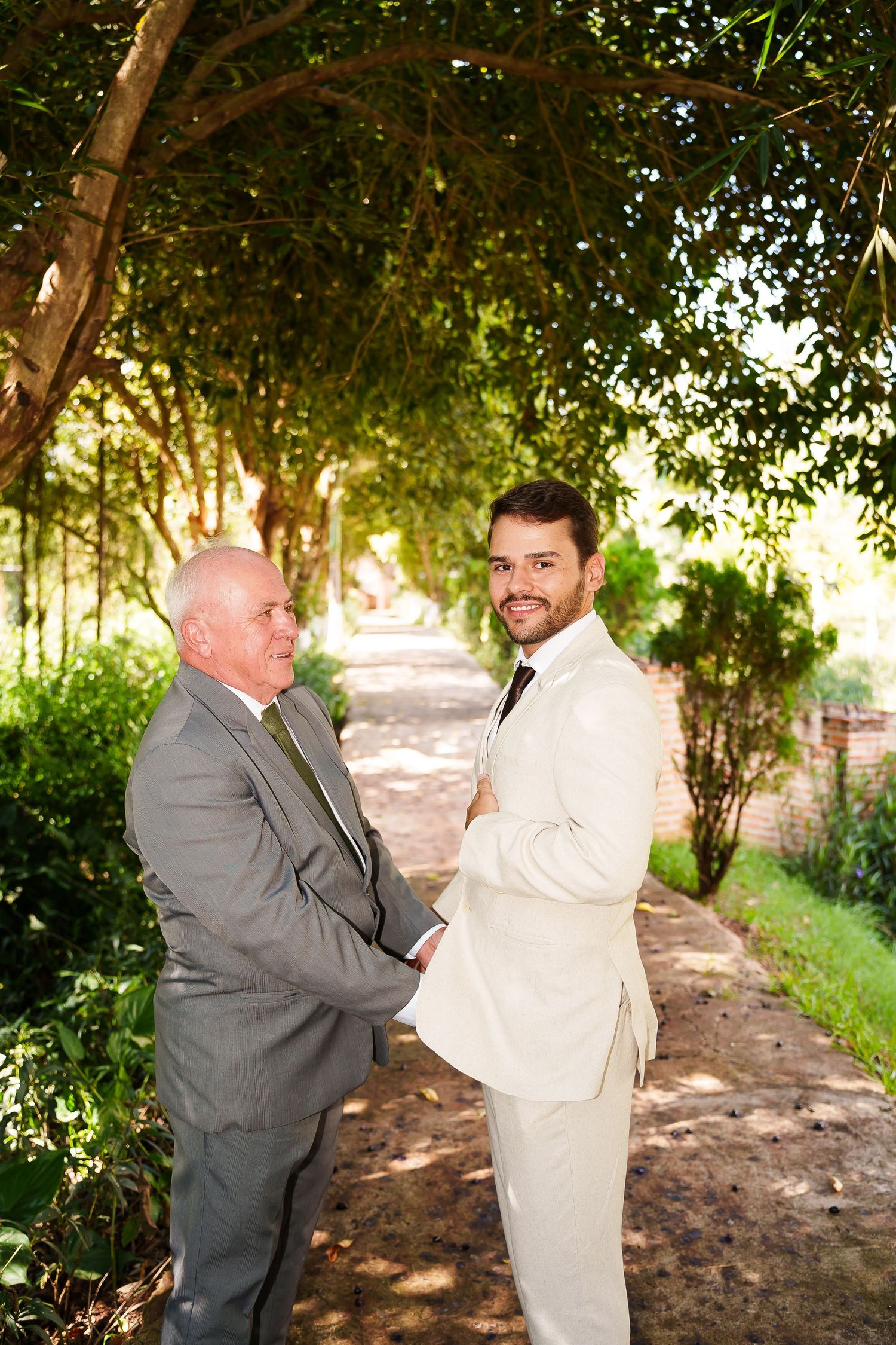 Two men in suits pose together outdoors under lush green trees on a sunny day at a wedding ceremony.