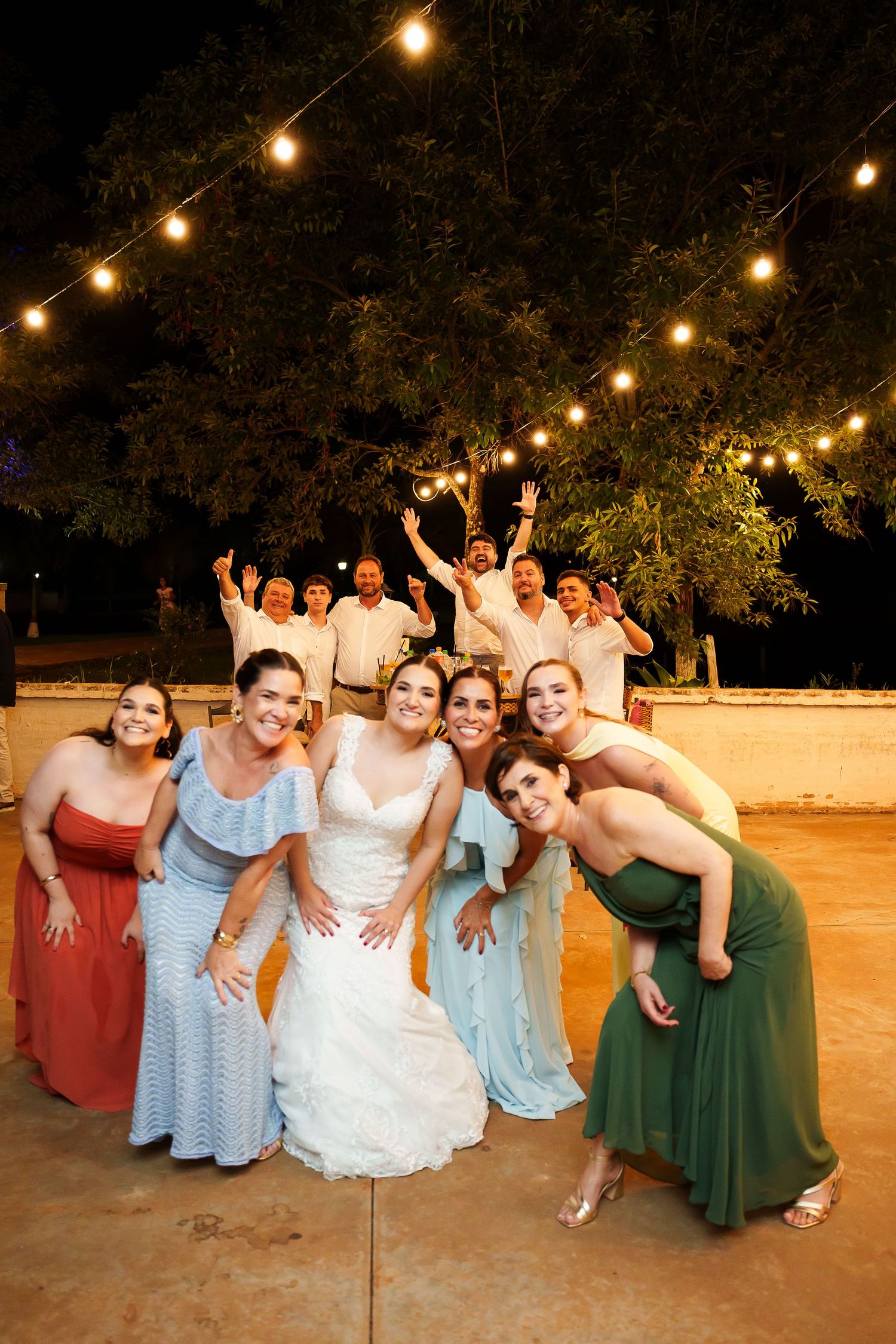 Bride poses with bridesmaids in colorful gowns and groomsmen celebrating outdoors at night under string lights.