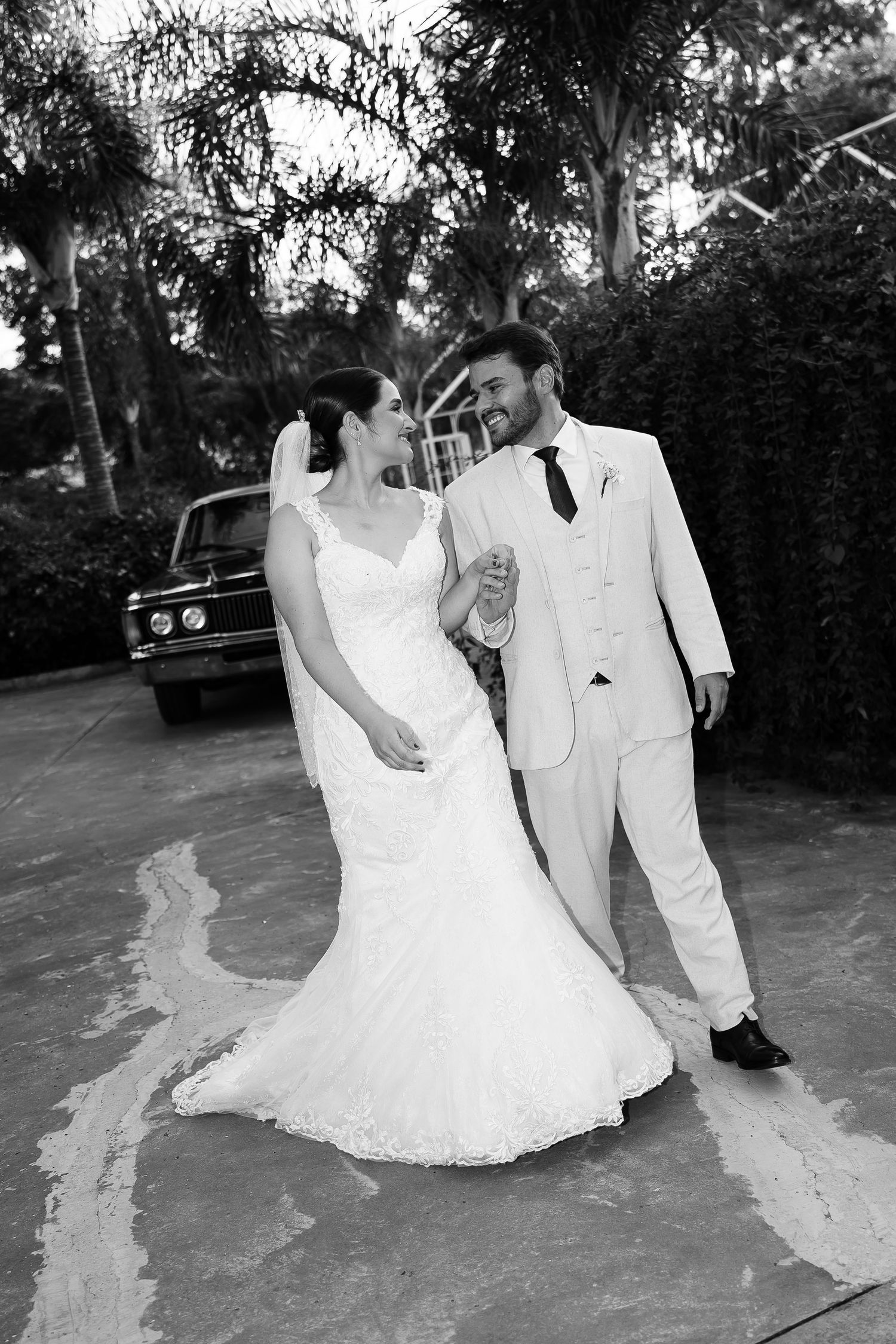 Black and white photo of a bride and groom walking together, smiling, with a vintage car and lush greenery in the background.
