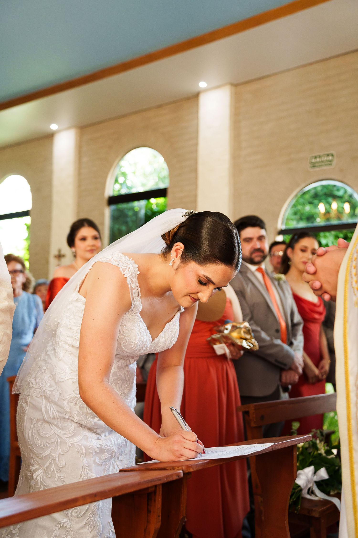 Bride in white lace gown signing marriage certificate at church altar during wedding ceremony.