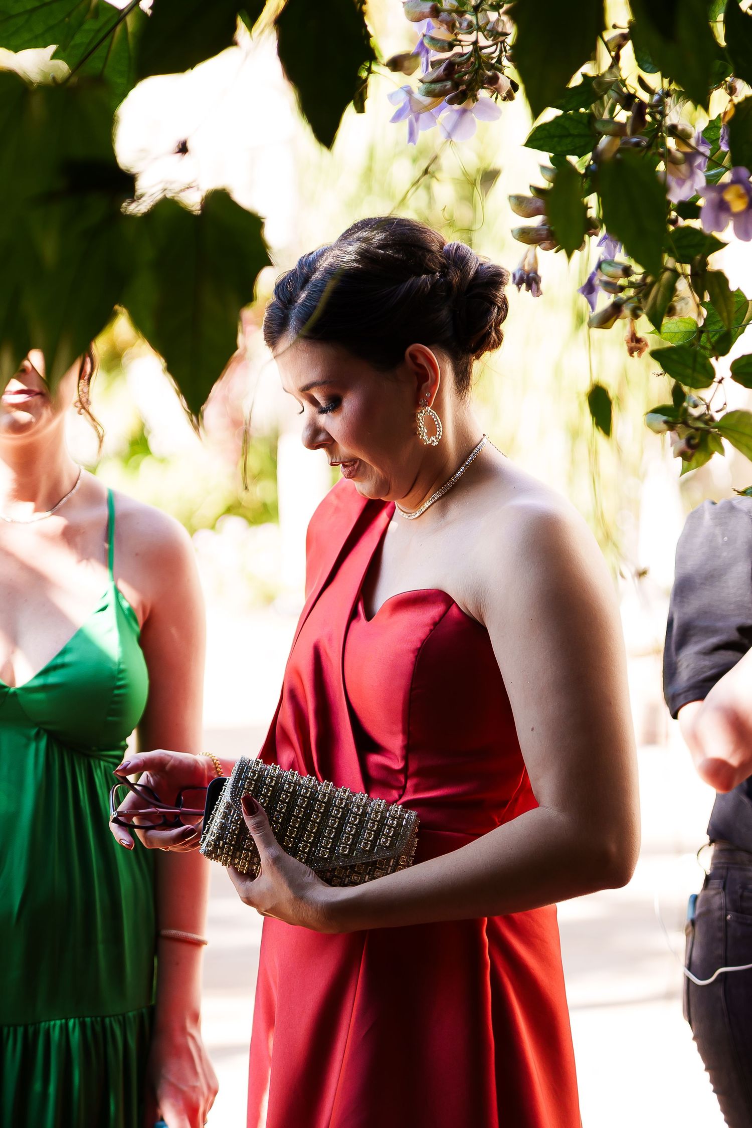 Woman in elegant red gown holding clutch purse, standing outdoors under green foliage at a formal event.