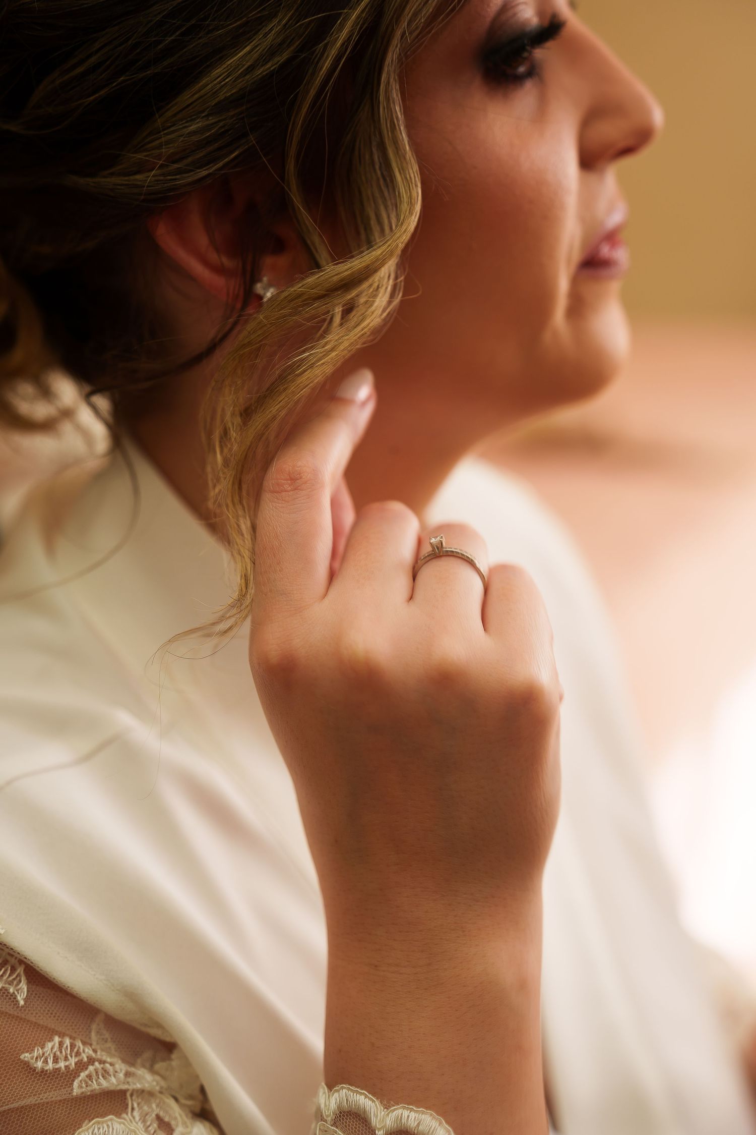 Bride touching her ear, showcasing a diamond engagement ring on her finger, wearing a white lace wedding dress.