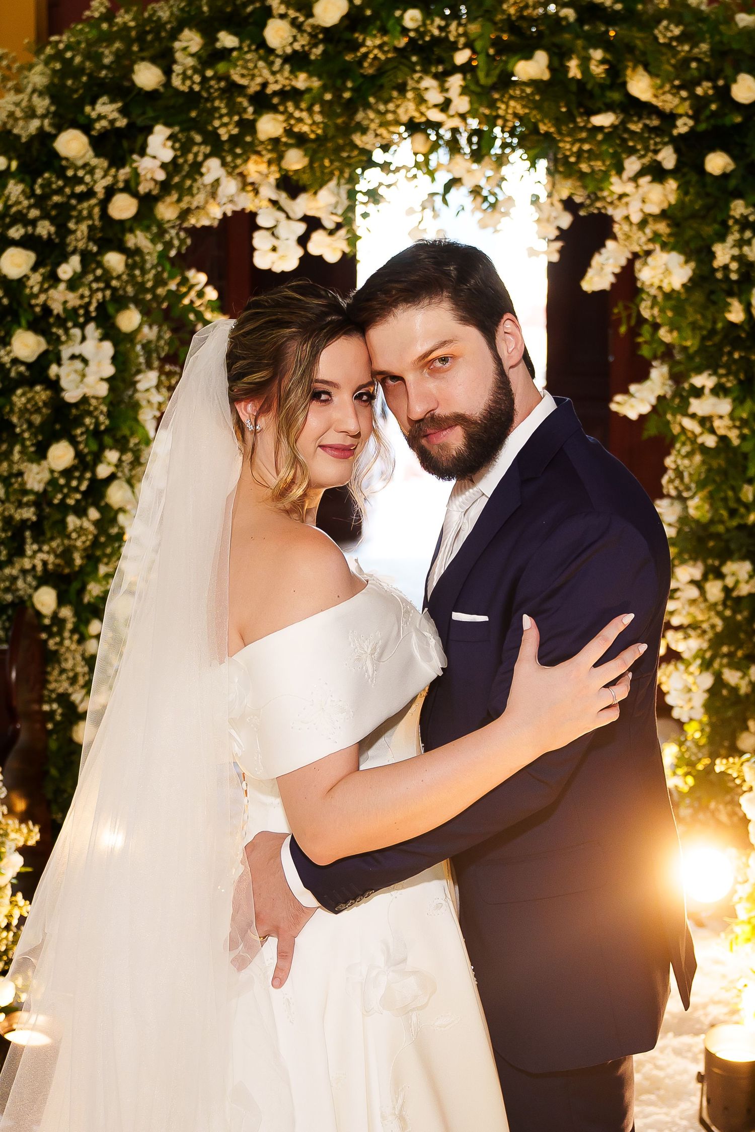 A bride and groom embrace tenderly under a floral arch adorned with white roses at their elegant wedding ceremony.