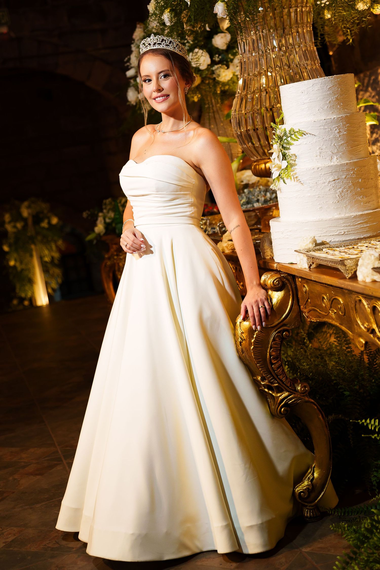 Bride in strapless ivory ball gown with tiara poses beside elegant white wedding cake at reception.