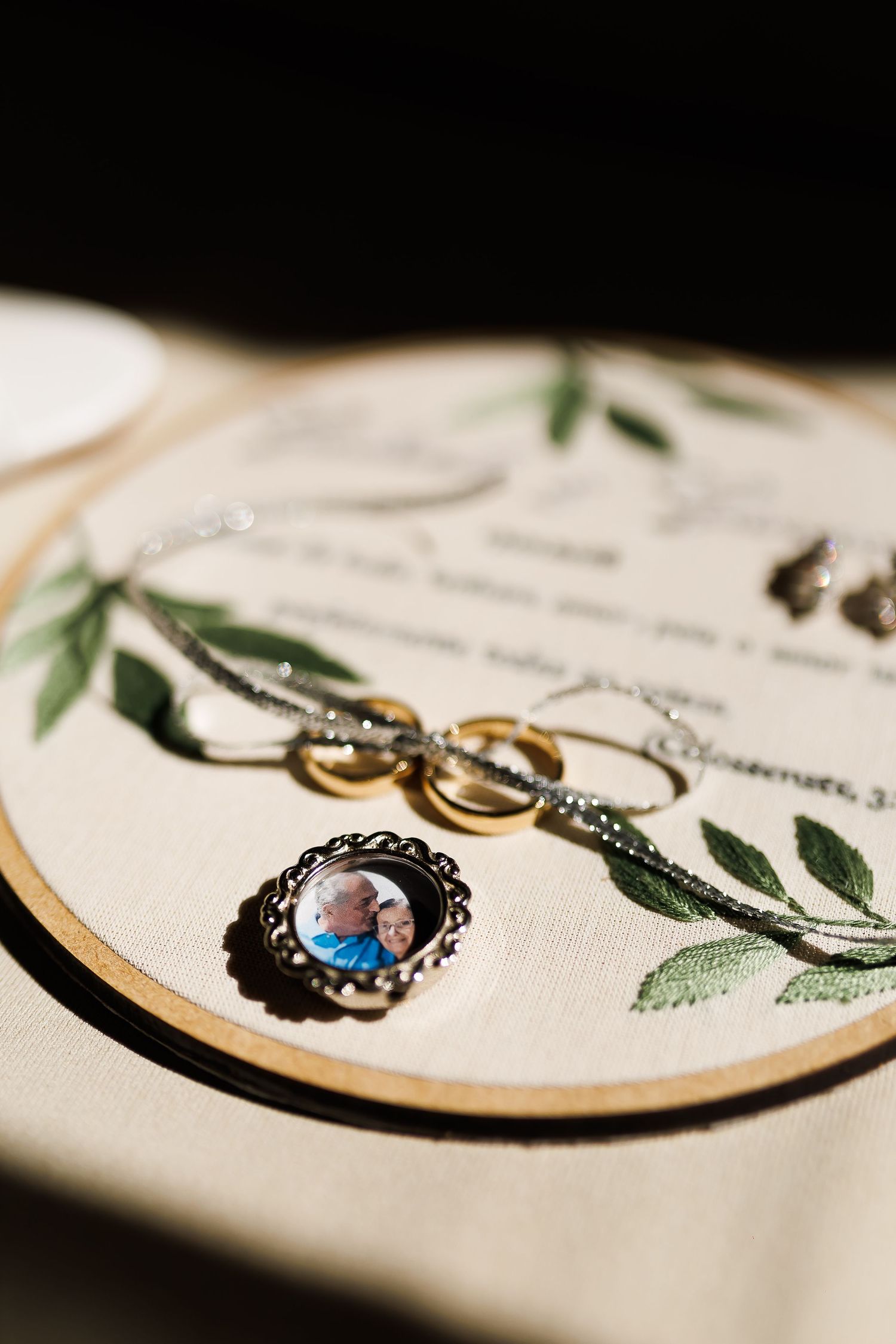 Wedding rings and a photo charm brooch displayed on an embroidered hoop with greenery and calligraphy details.