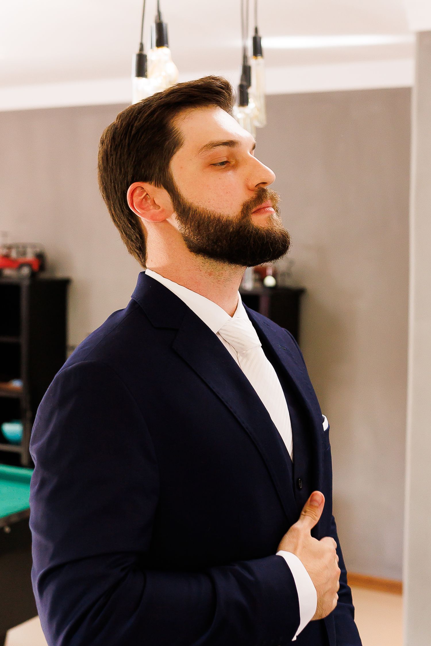 A bearded man in a navy blue suit adjusts his jacket lapel indoors under pendant lights.