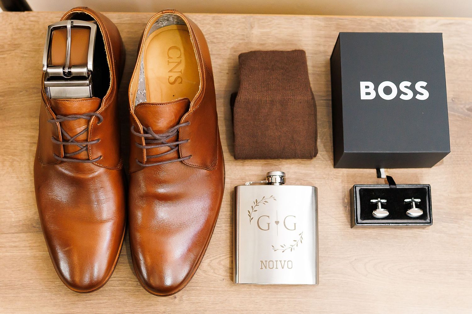 Brown leather dress shoes, belt, socks, engraved flask, cufflinks, and Hugo Boss box arranged as groom wedding accessories.