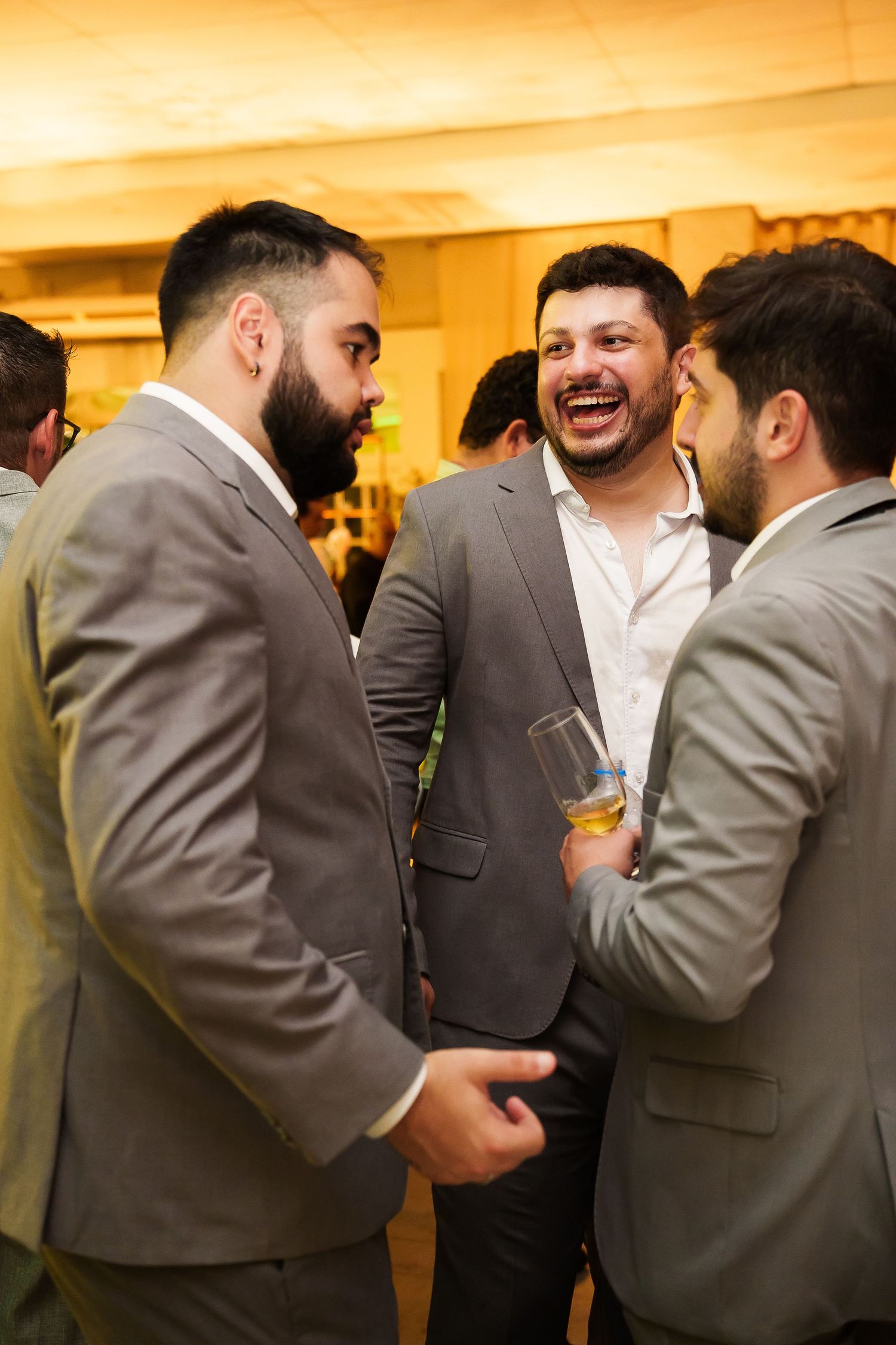 Three men in gray suits laughing and socializing at a formal event, one holding a glass of whiskey.