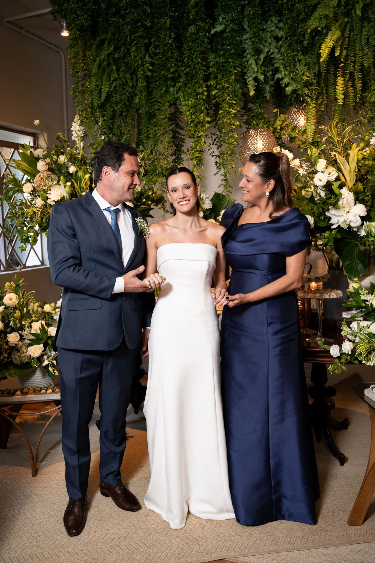 Bride in white strapless gown poses with man in navy suit and woman in navy dress at floral wedding ceremony.