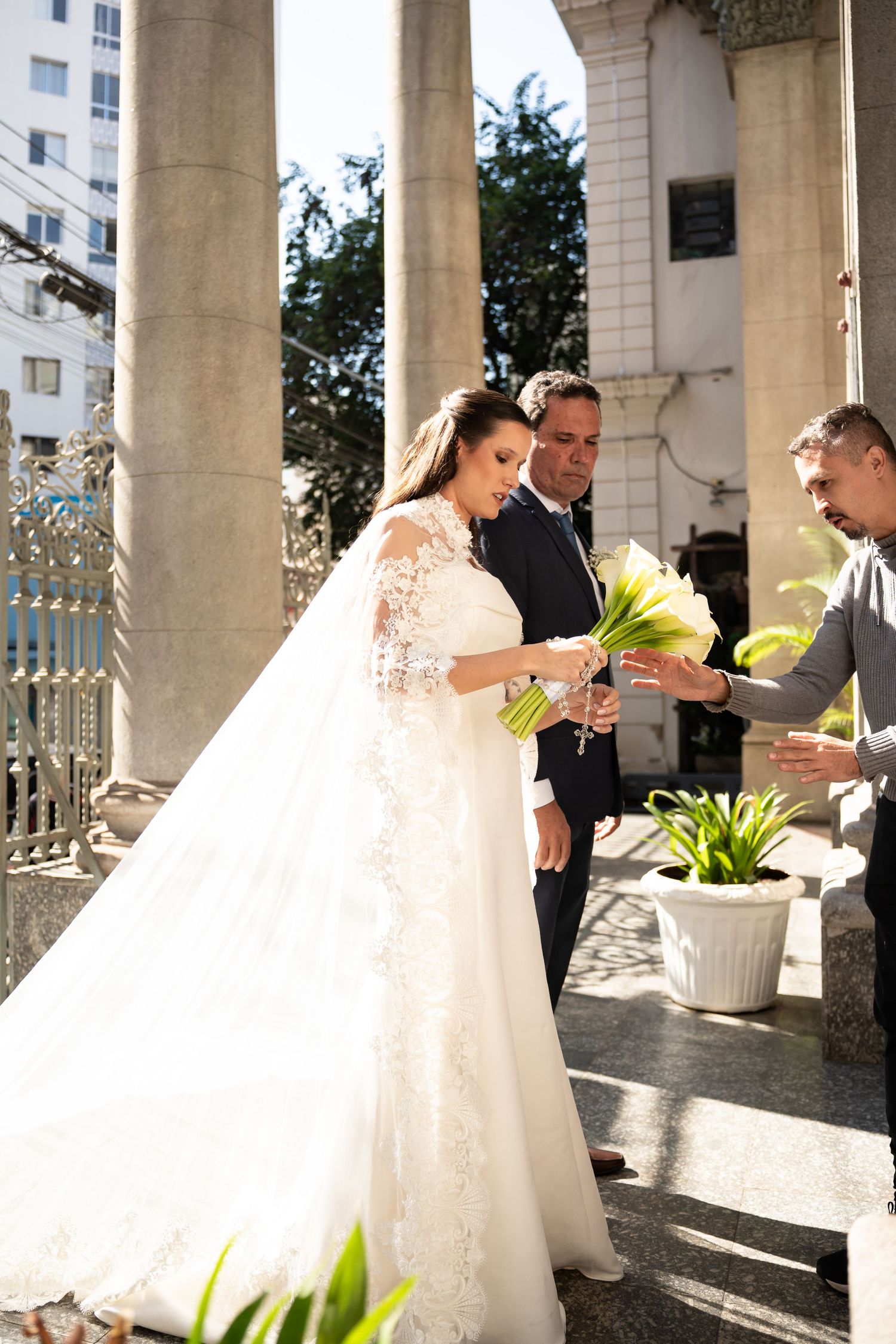 Bride in white lace gown and veil receives bouquet of white flowers from guest outside church columns.