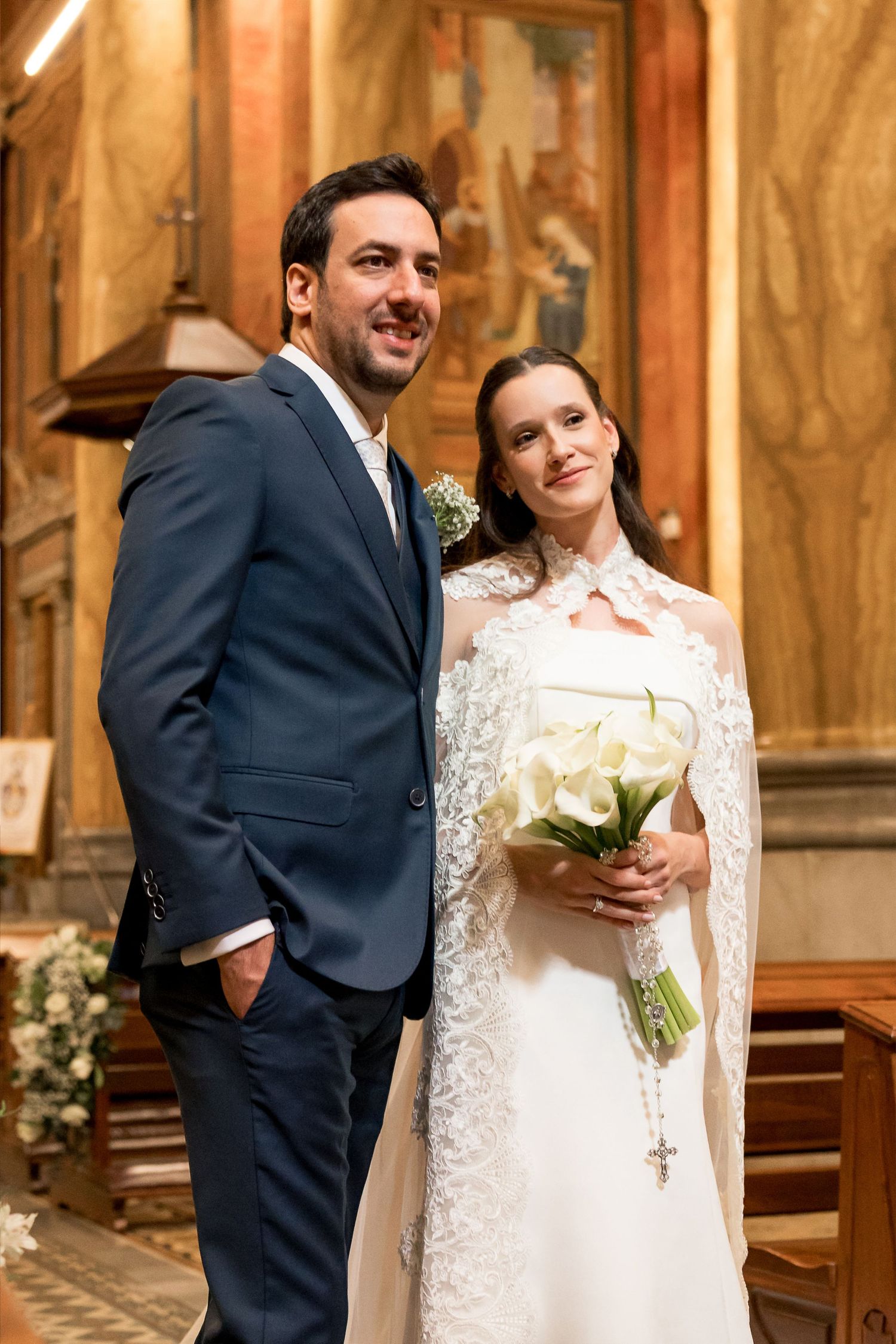 Bride in lace cape holding white calla lily bouquet stands with groom in navy suit inside a church.
