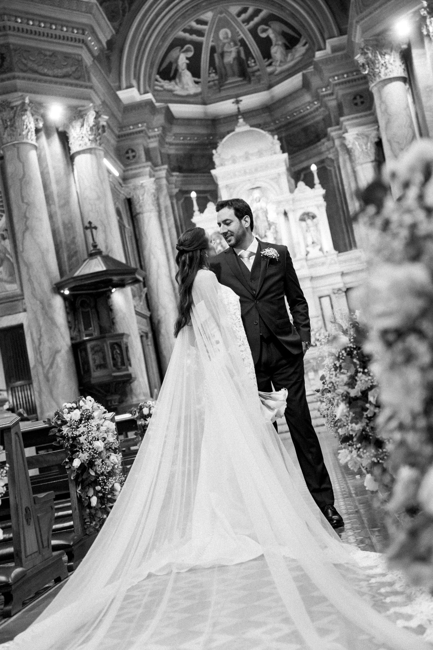 Black and white photo of bride and groom sharing a romantic moment inside an ornate church with floral arrangements.